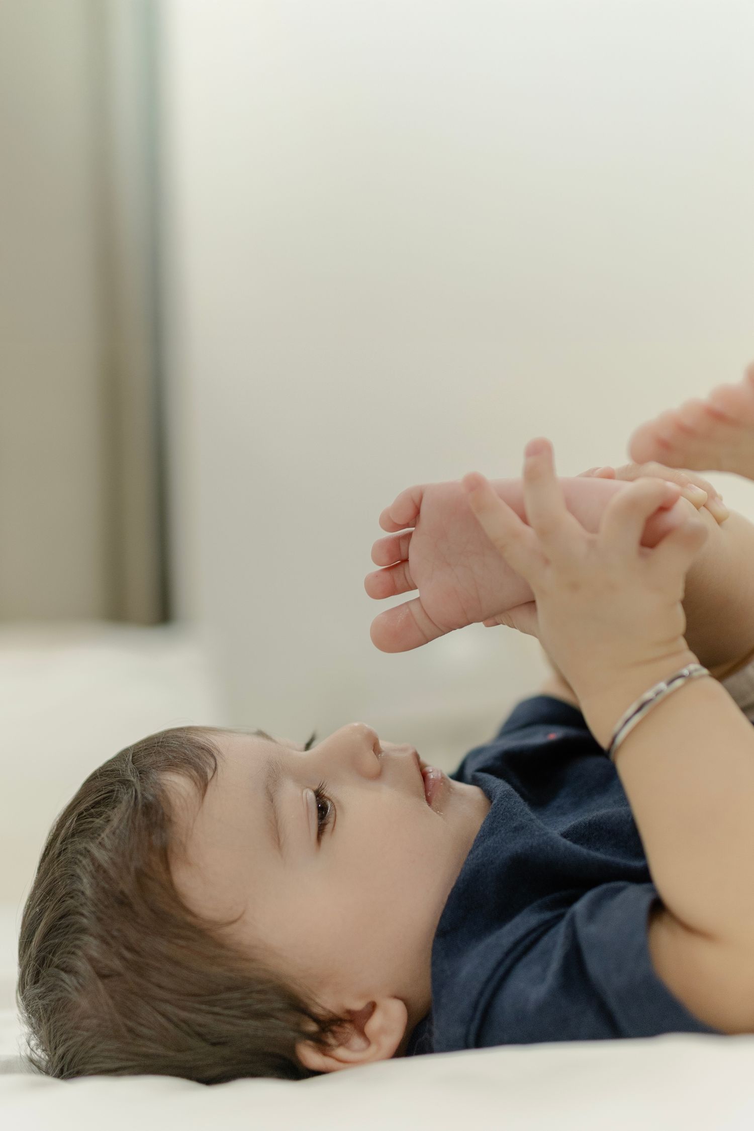 A young child lies on a white bed playing with their feet while wearing a navy blue shirt.