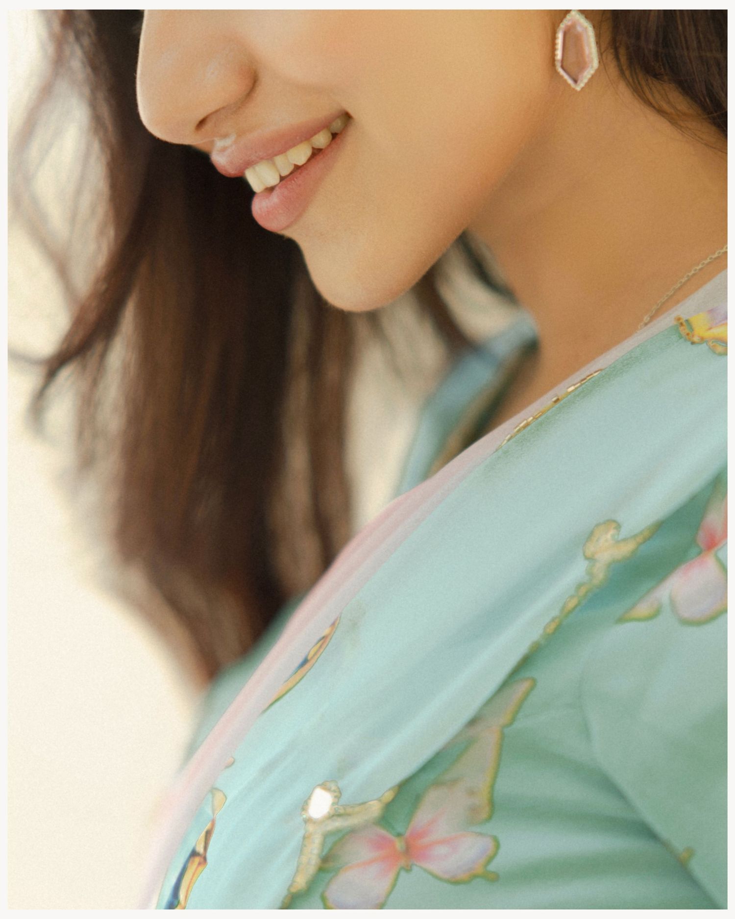 Close-up of smiling woman in teal floral outfit with pink butterfly embroidery and elegant earring.