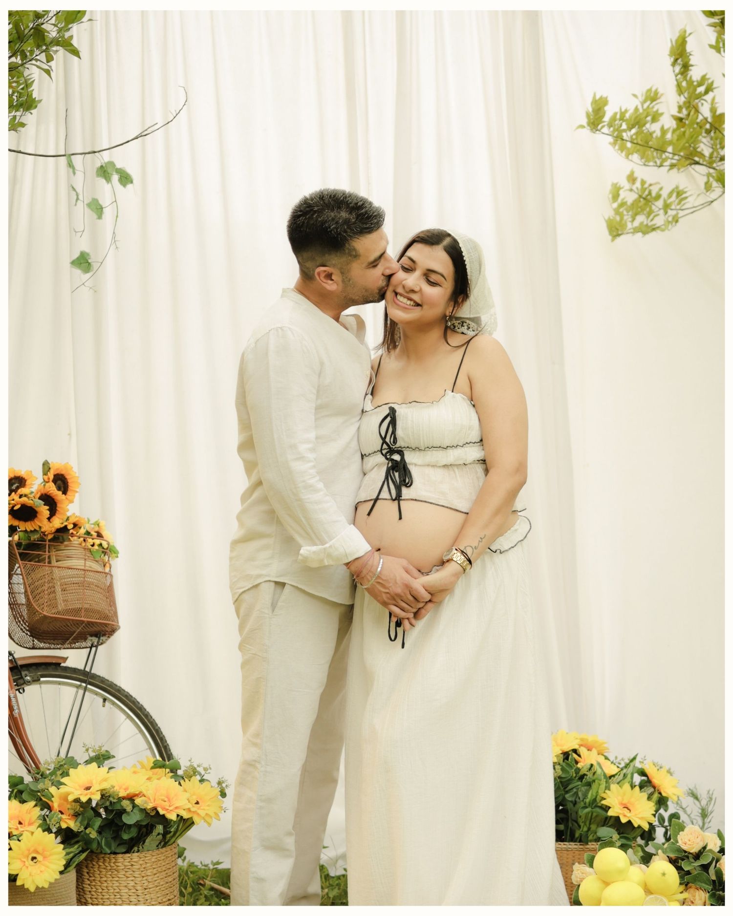 Couple poses for maternity shoot with sunflowers and white drape backdrop, man kissing pregnant woman's cheek.