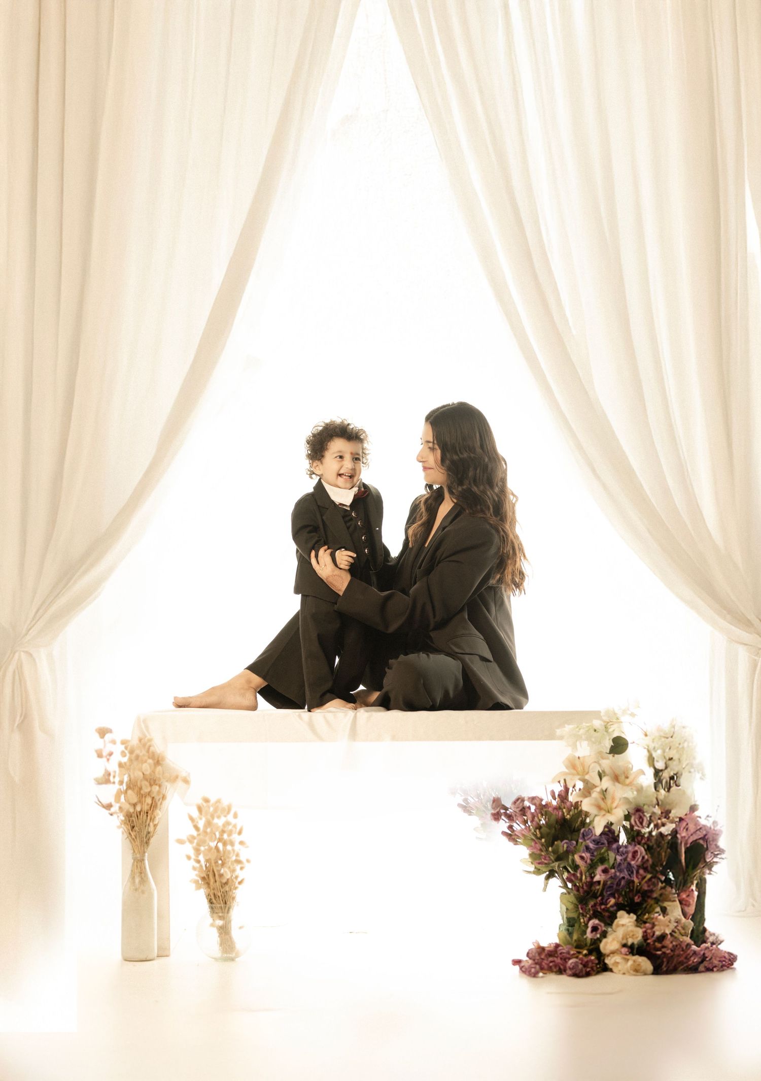 A woman and young child in matching black outfits sit together on a white surface surrounded by flowers and white curtains.