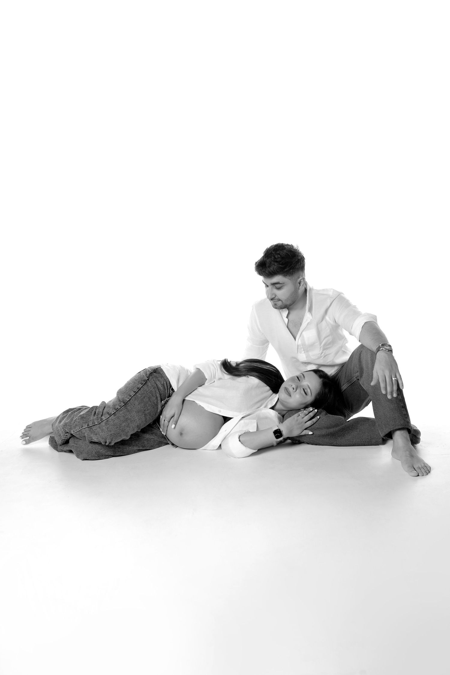 Black and white photo of a pregnant couple sitting on floor, woman resting head on mans lap in studio.