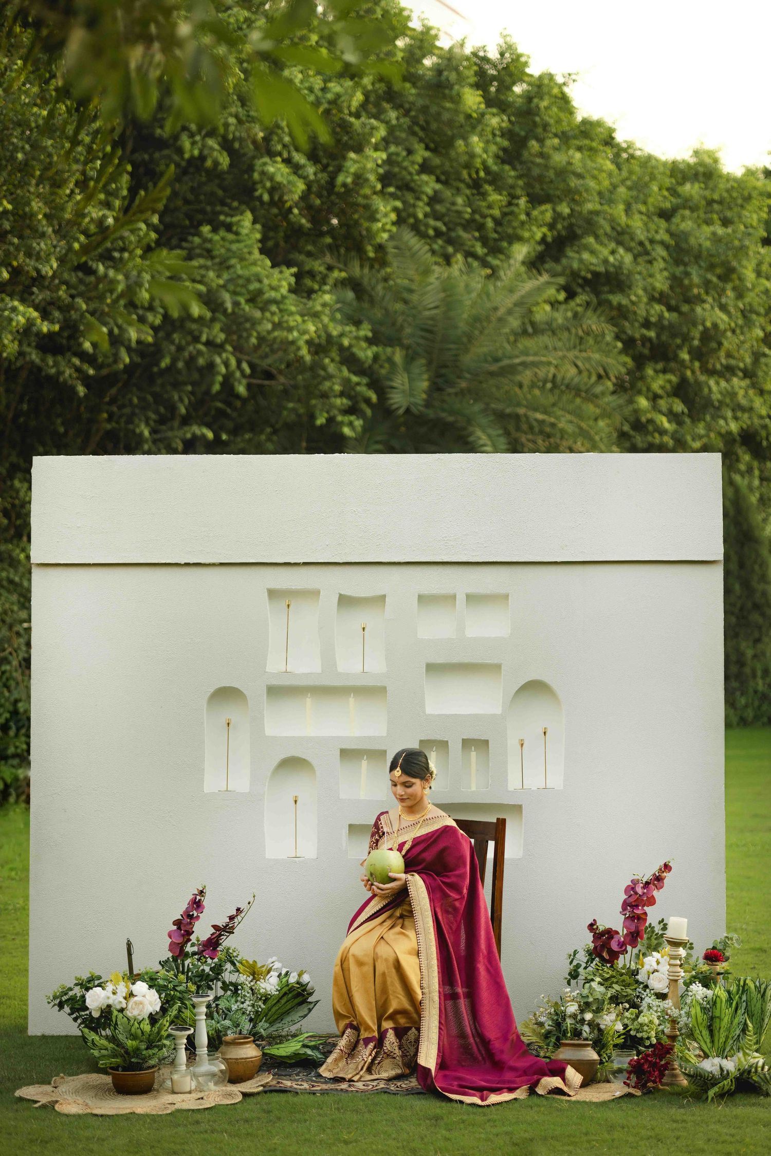 Woman in elegant maroon and gold saree seated before white decorative backdrop with floral arrangements outdoors.
