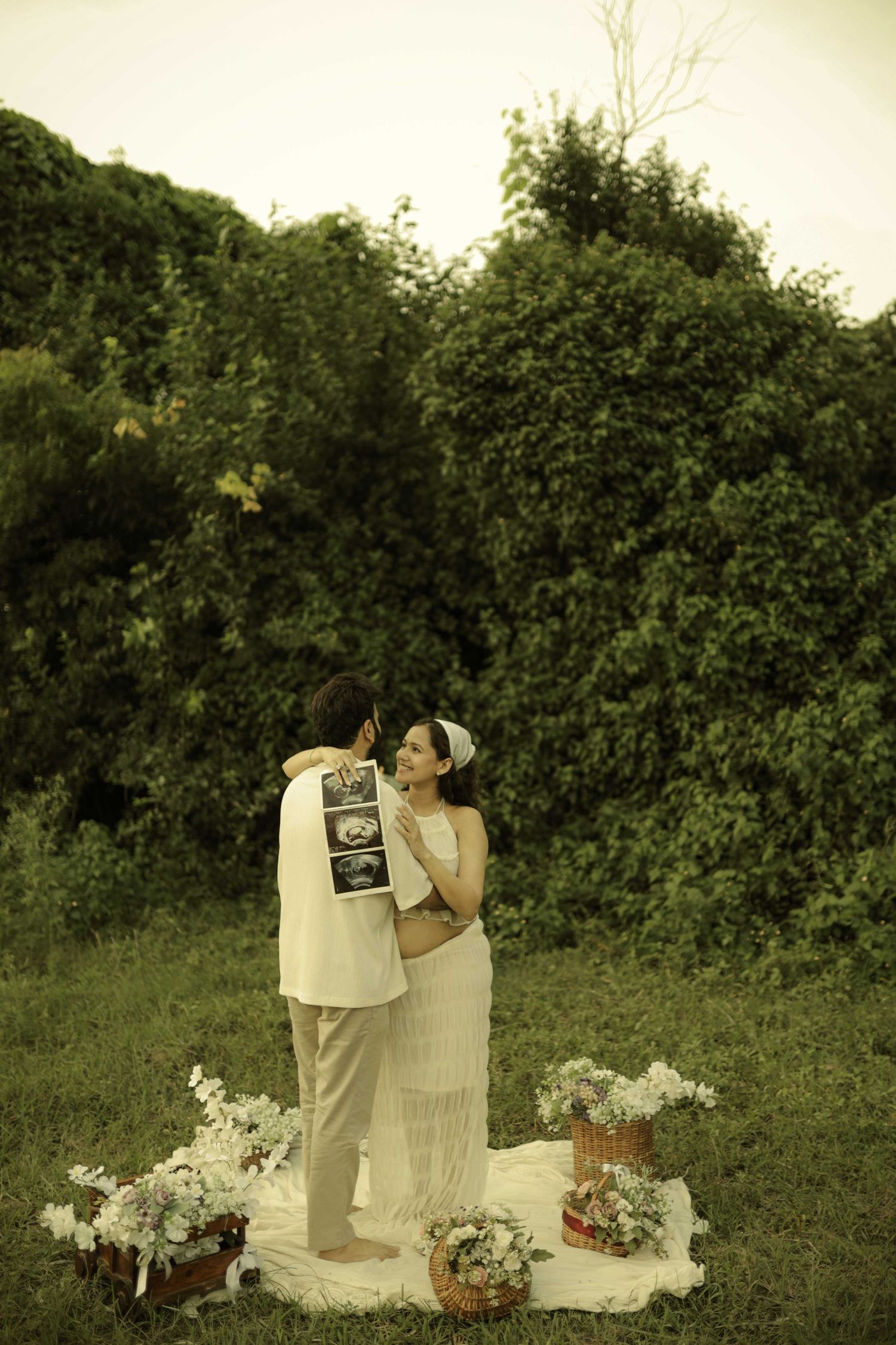Couple sharing tender moment outdoors, holding ultrasound photos, surrounded by flowers on a blanket.