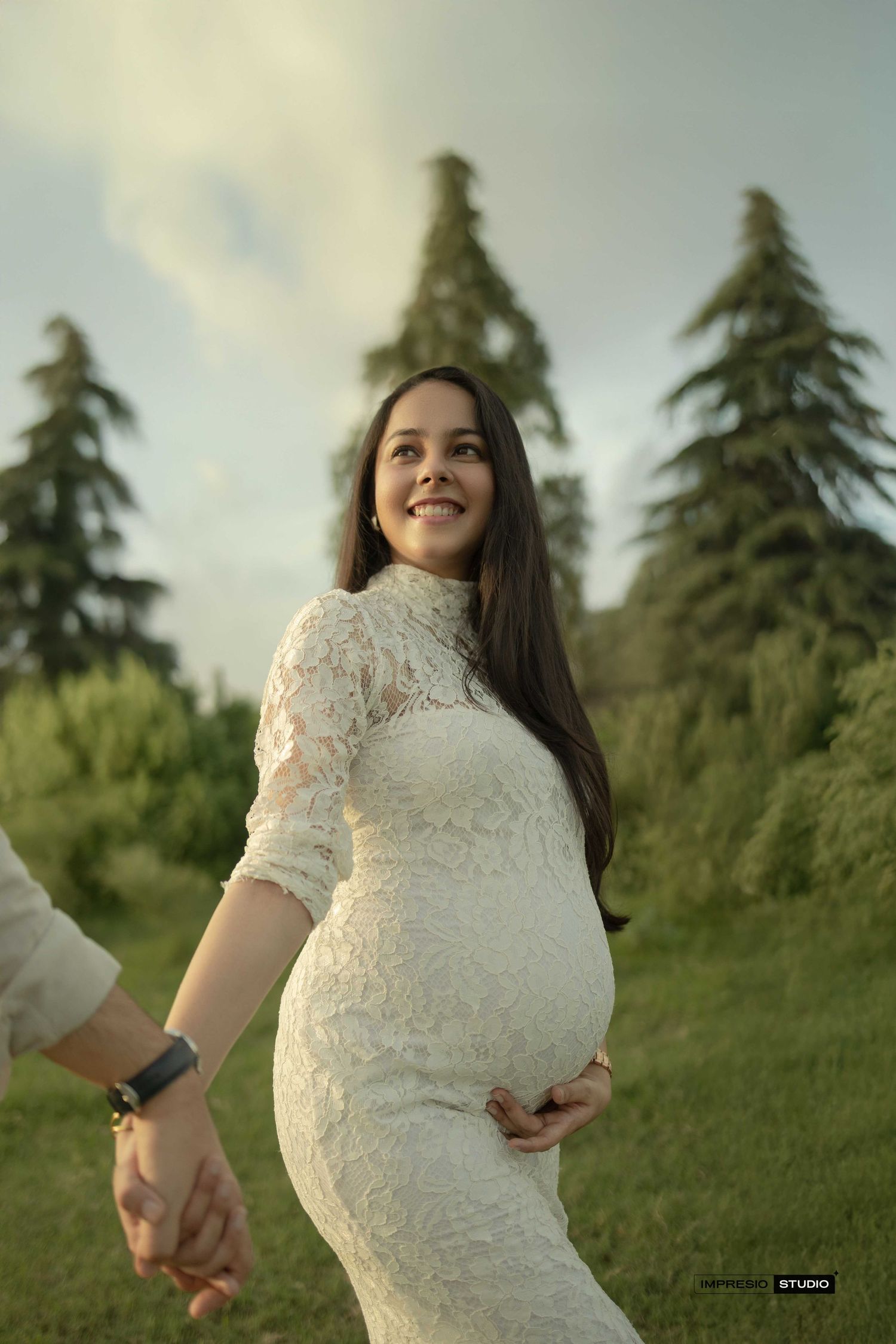 Smiling pregnant woman in white lace dress holds partner's hand outdoors among tall evergreen trees.