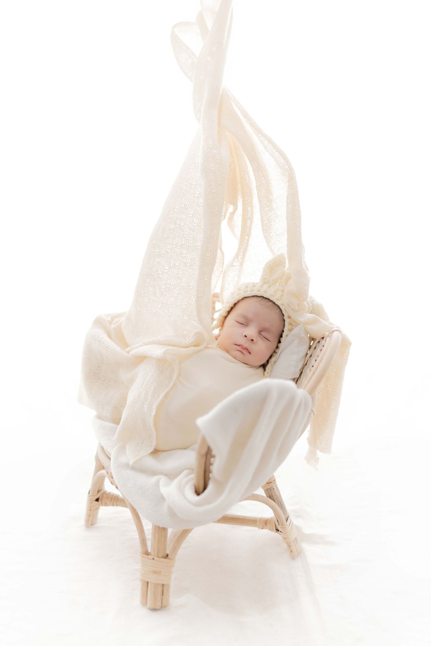 Peaceful newborn baby sleeping in small wicker chair wrapped in cream blanket with knit bonnet.