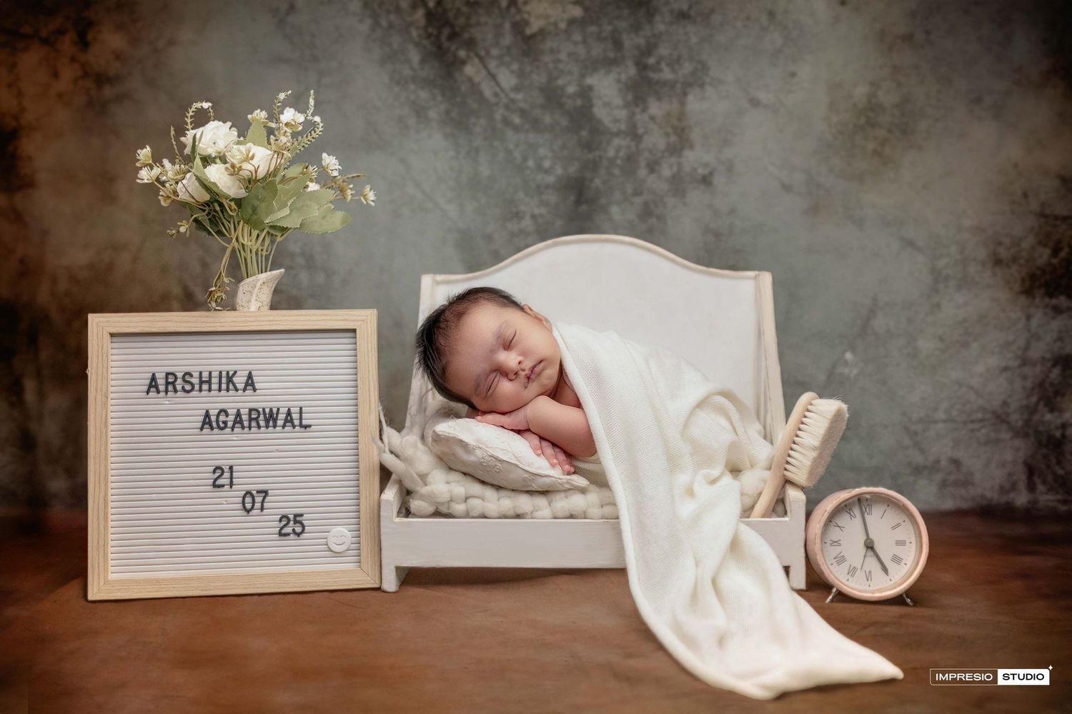 Newborn baby Arshika Agarwal sleeping peacefully on a white mini bed with a letter board and clock in a studio.