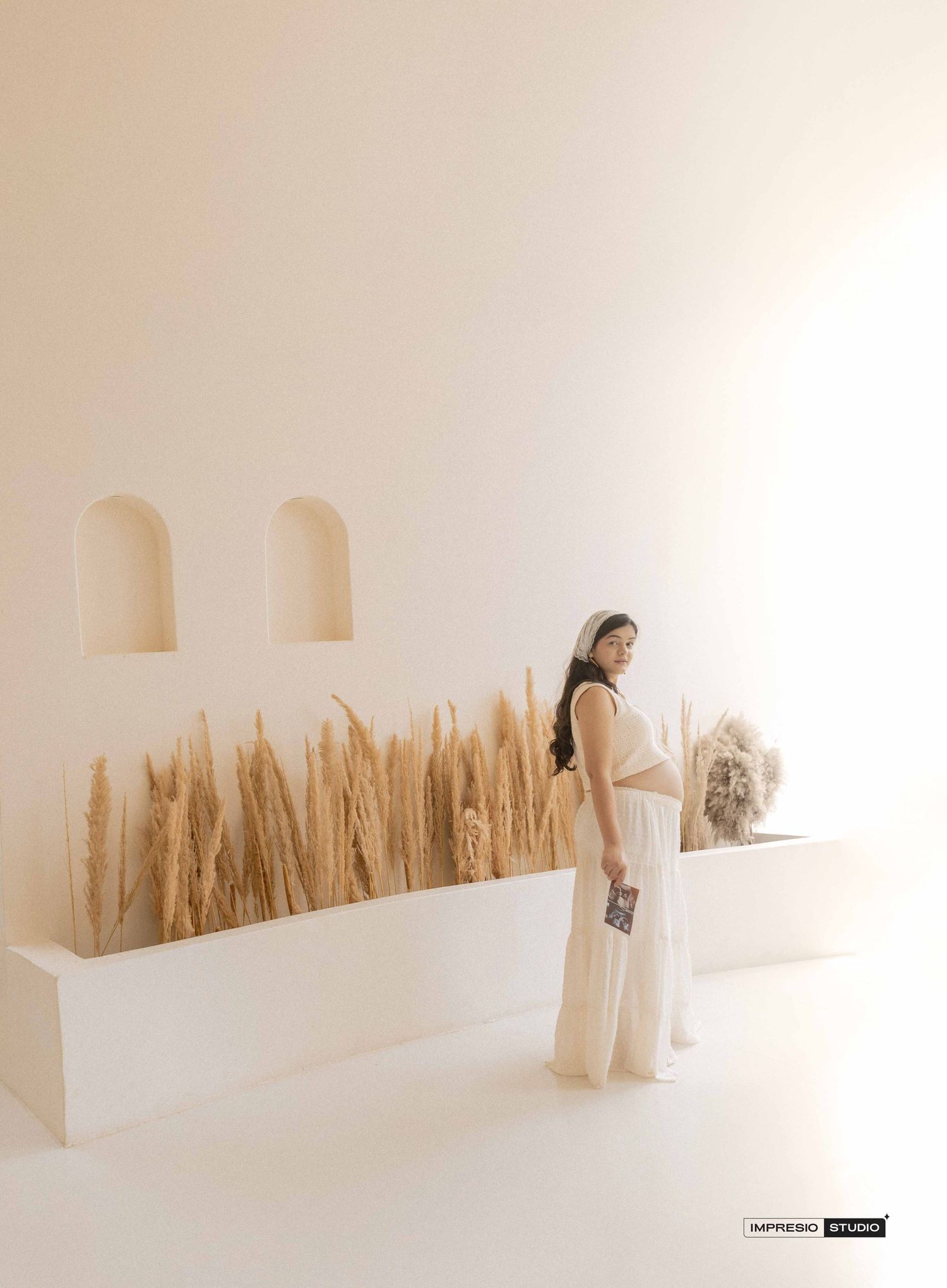Woman in white two-piece outfit poses beside dried pampas grass display in minimalist white studio setting.