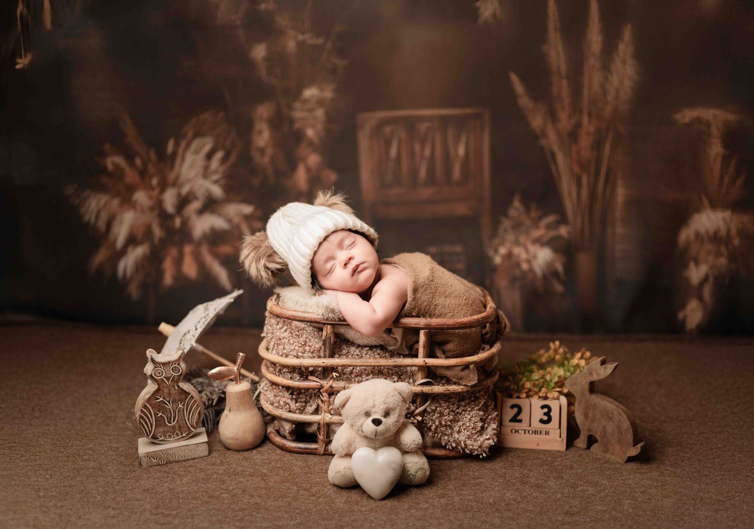 Newborn baby sleeping in rustic wooden basket with teddy bear, white knit hat, and vintage props in professional studio photo.