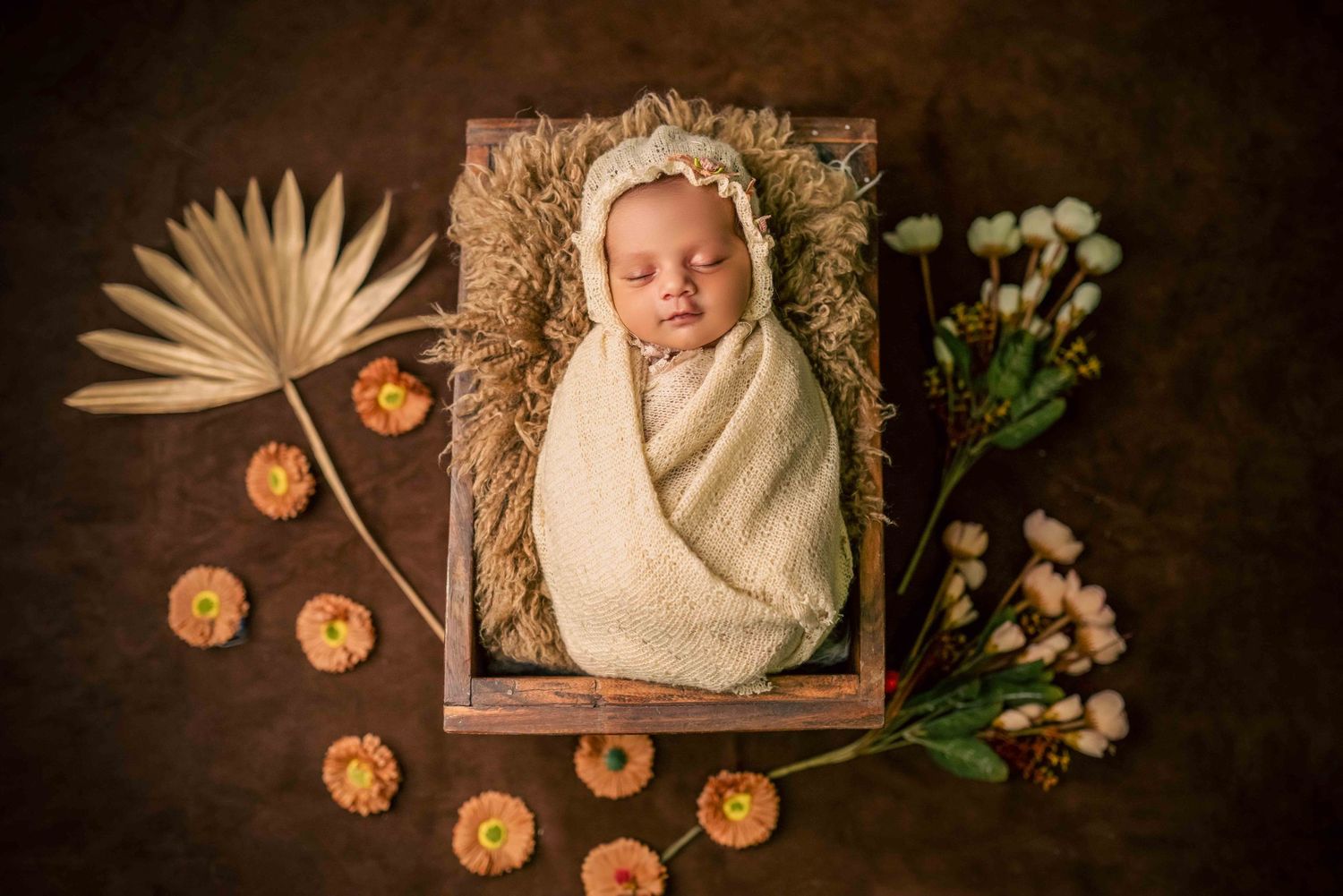 Newborn baby swaddled in cream wrap sleeping in wooden box with floral decorations and dried palm leaf.