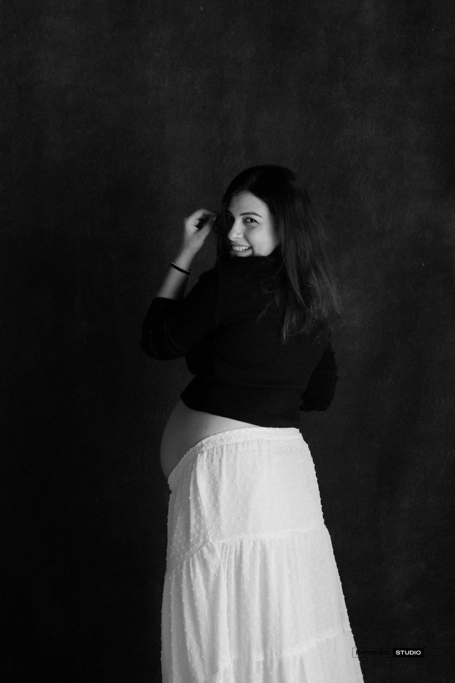 Black and white studio portrait of a pregnant woman in black turtleneck and white skirt, smiling over her shoulder.
