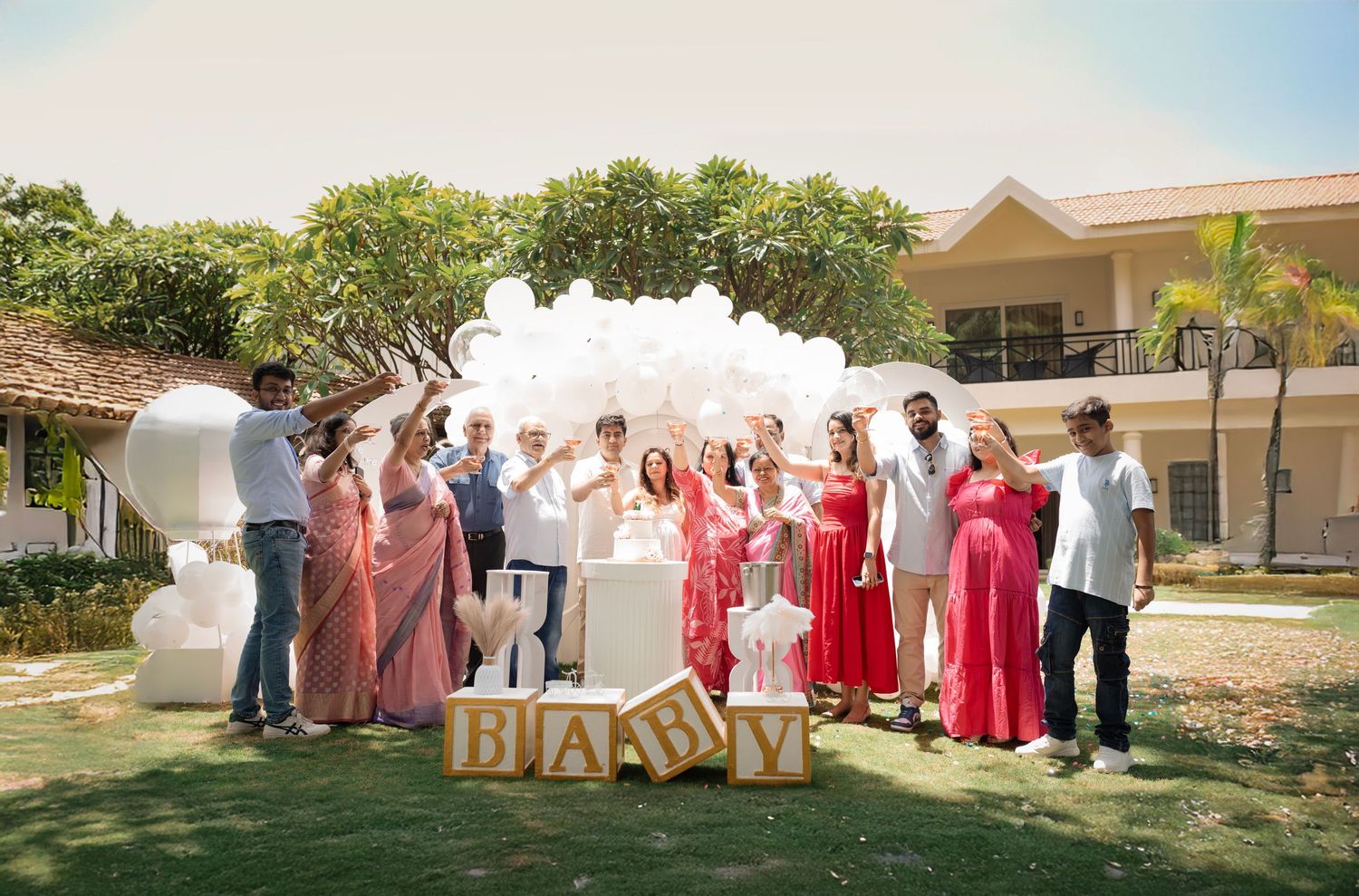 Group celebrating a baby shower outdoors with white balloons, BABY blocks, and a cake on a sunny day.