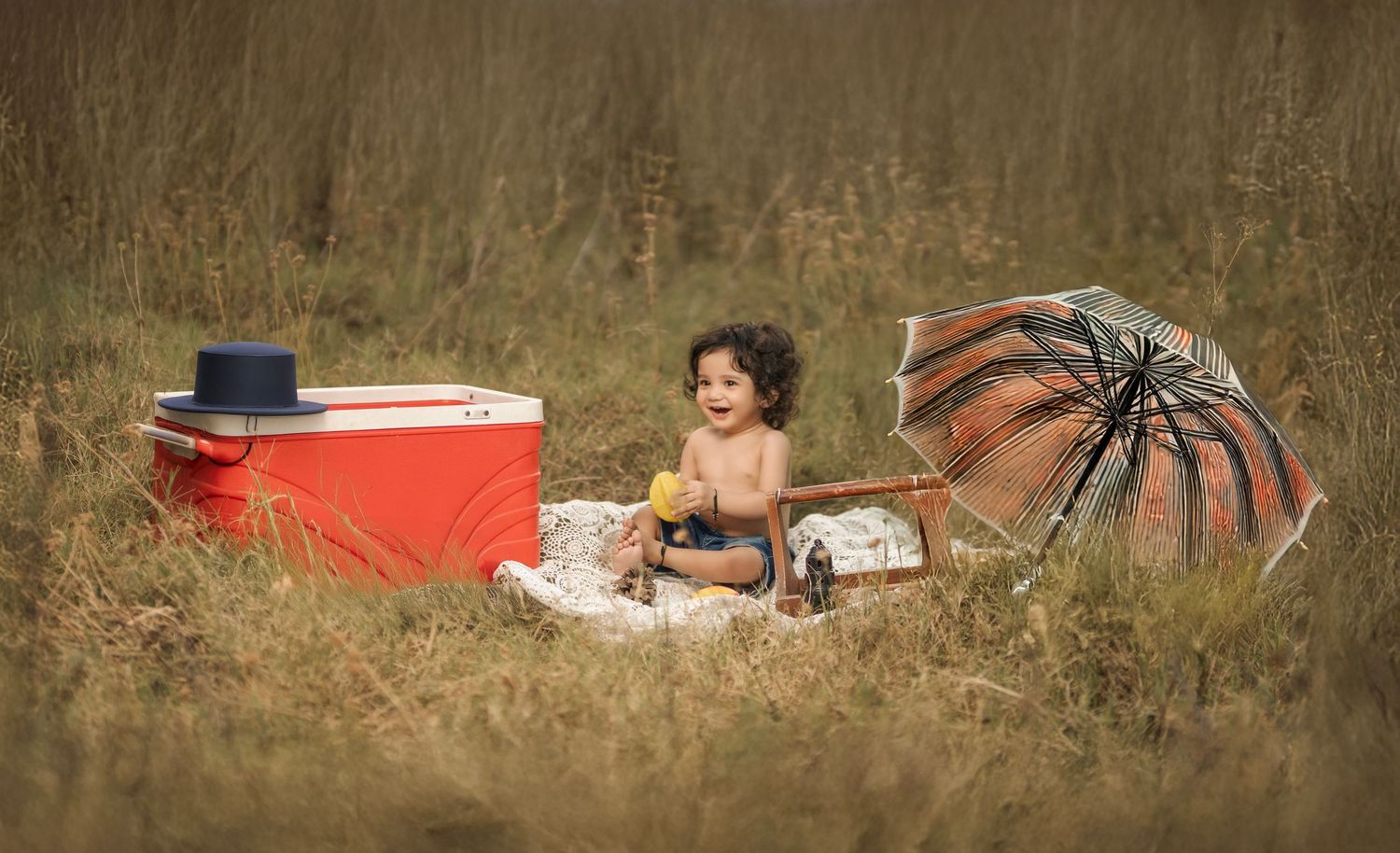 A toddler sits outdoors in tall grass surrounded by a red cooler, hat, and open umbrella during a picnic.
