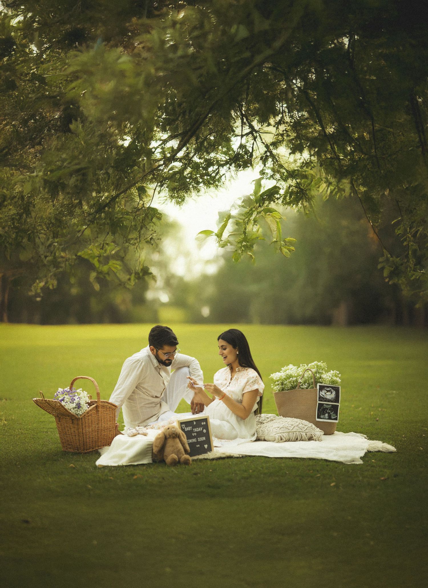 A couple lounges on a white blanket under trees with a picnic basket in a serene garden setting at sunset.