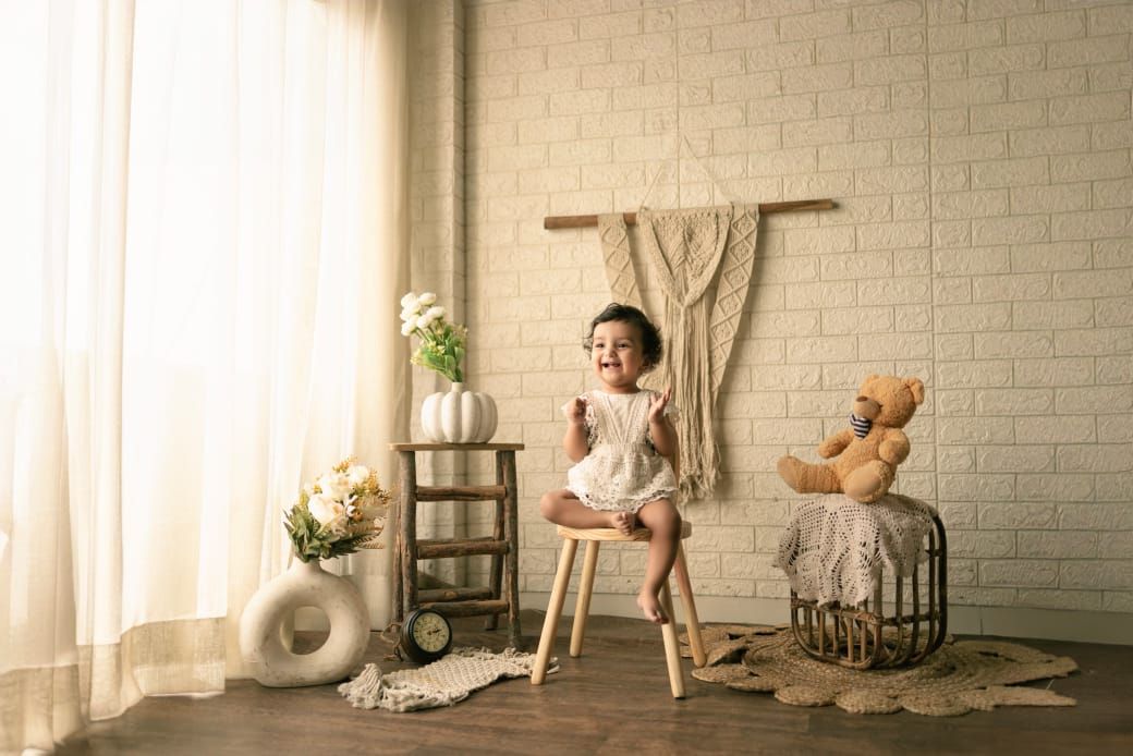 Vintage style photo with a child on stool next to teddy bear and decor against white brick wall pre birthday photoshoot