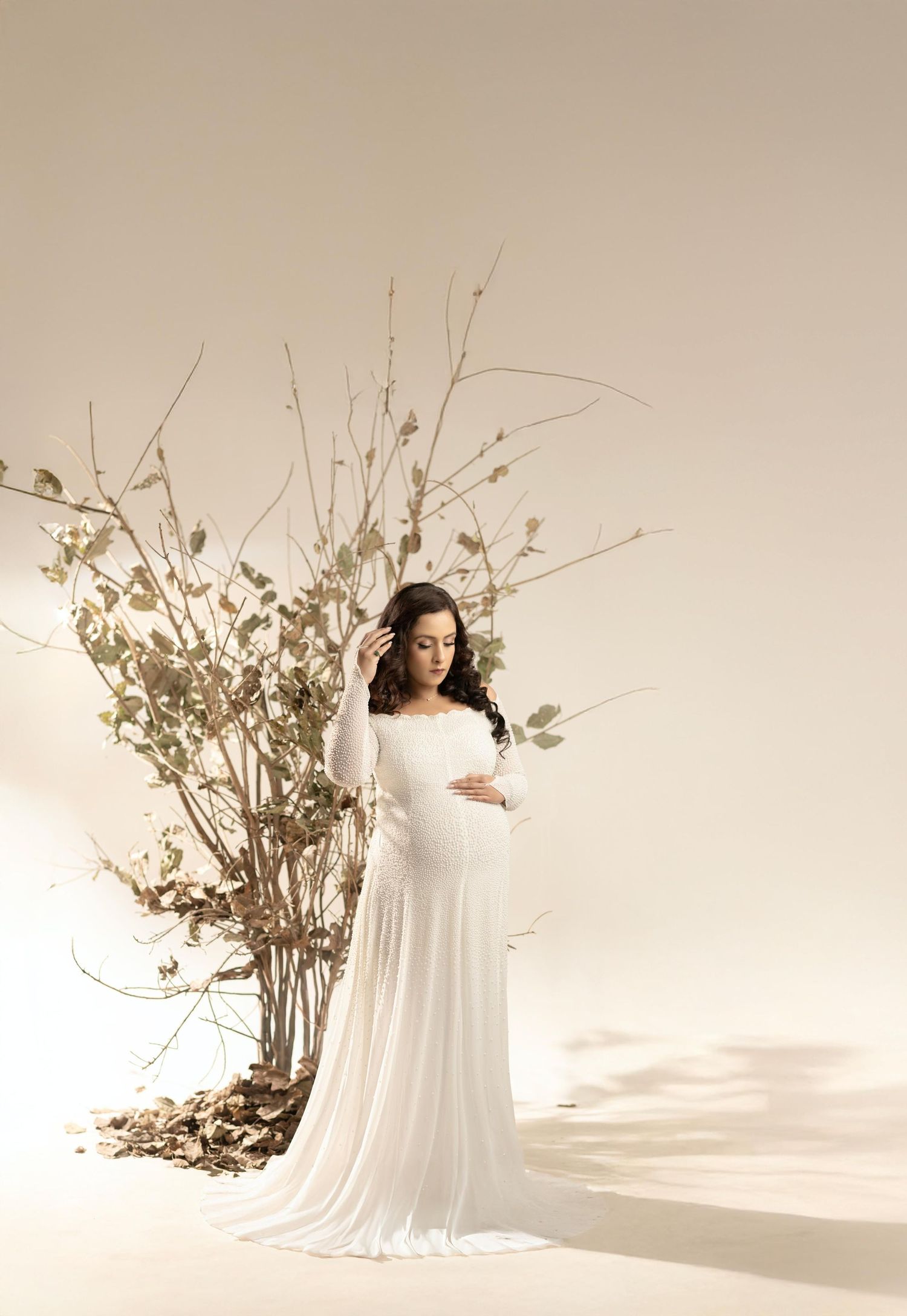 Artistic maternity photo shoot in white gown with dried floral branches against neutral backdrop.