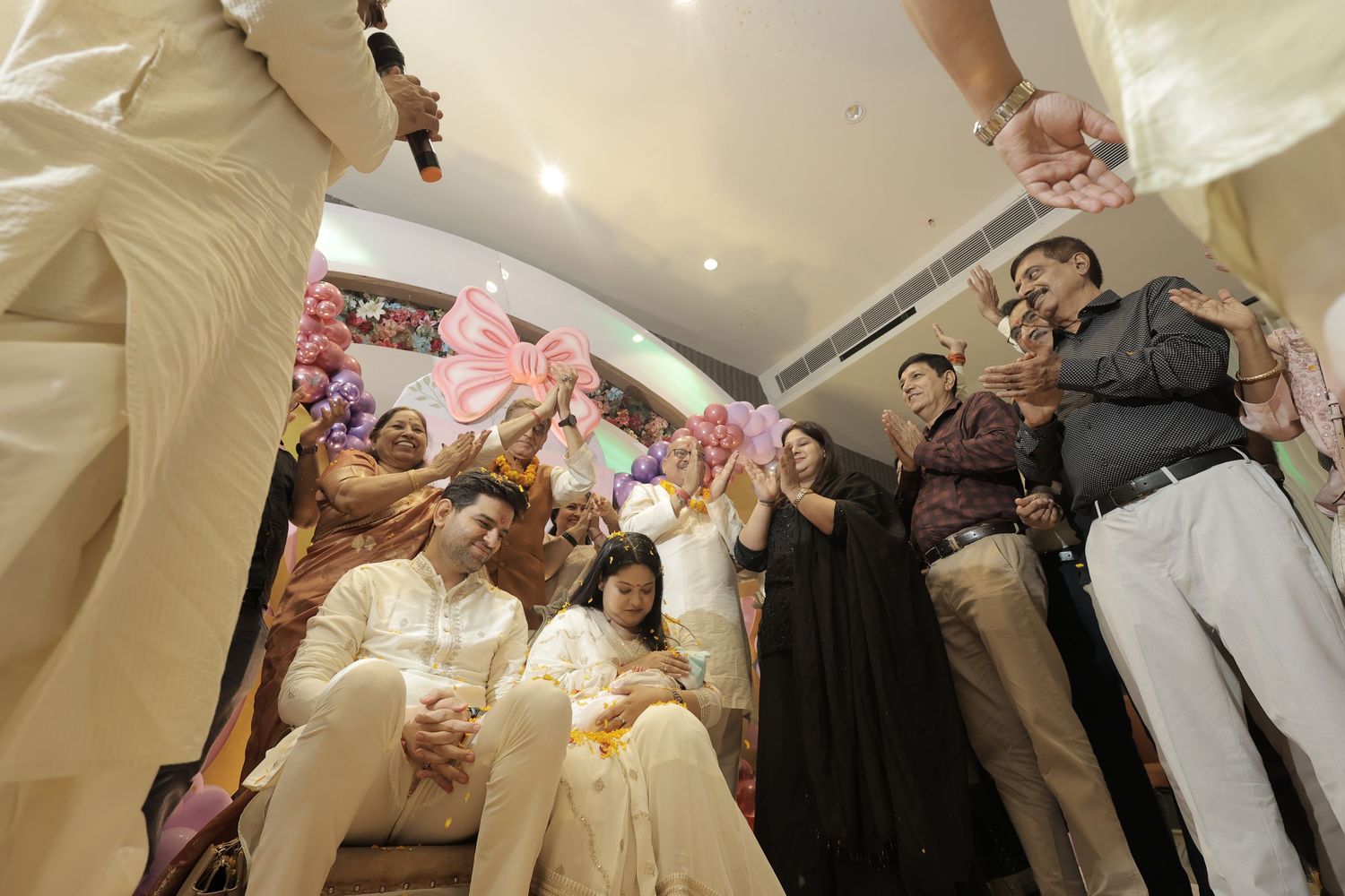 A seated couple in cream traditional attire is surrounded by smiling guests during a festive ceremony with pink floral decorations.