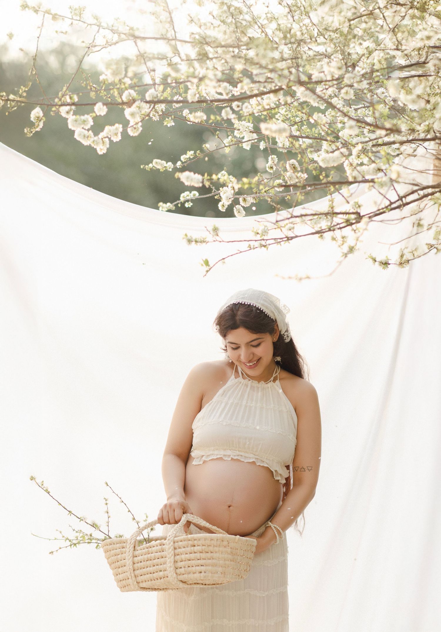 Pregnant woman in white crop top holding a wicker basket under blooming cherry blossom tree outdoors.