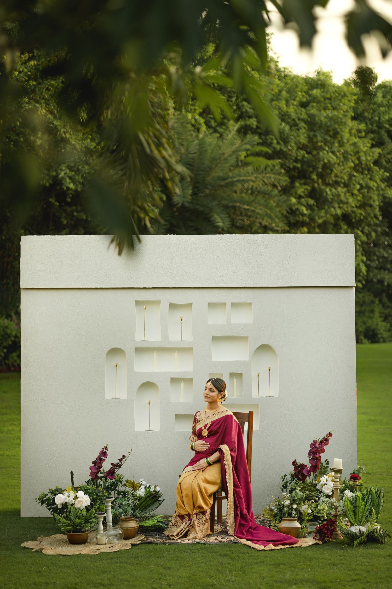 A bride in a maroon and gold saree sits gracefully before a white decorative backdrop with floral arrangements outdoors.