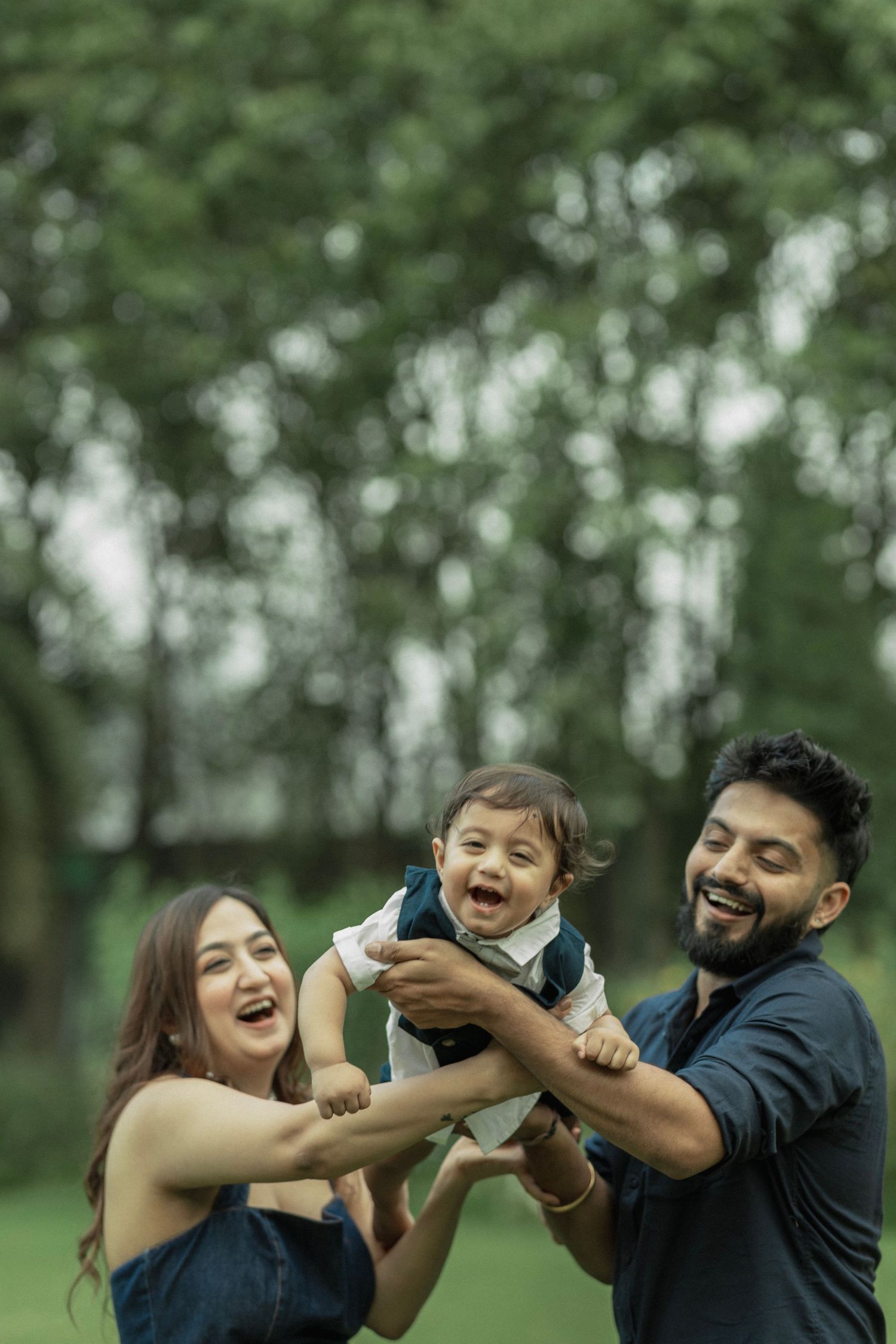 Happy Indian family with parents lifting laughing toddler in a lush green park outdoor setting.