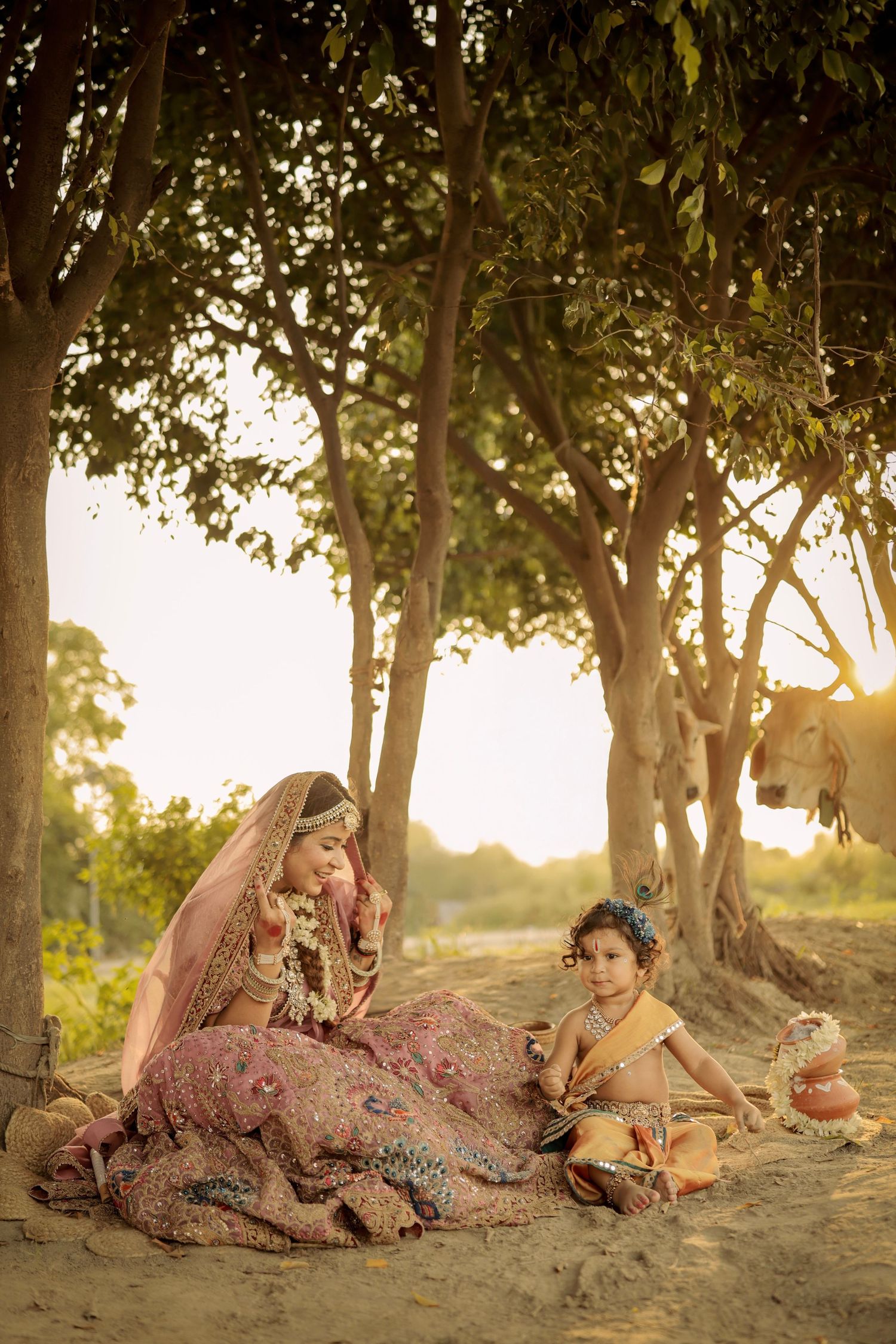 Indian bride in pink lehenga sits with child dressed as Krishna under golden trees at sunset.