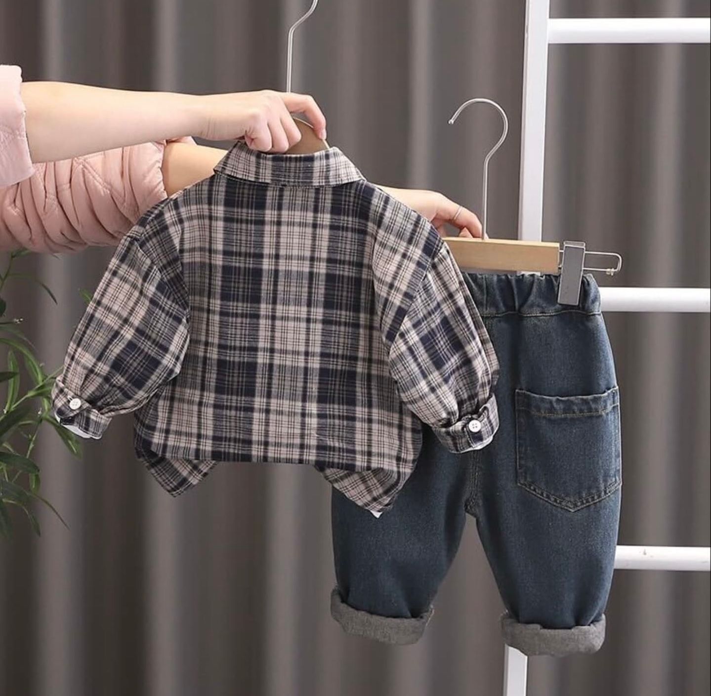 Hands hold a small pair of denim overalls on a white wooden ladder next to gray curtains.