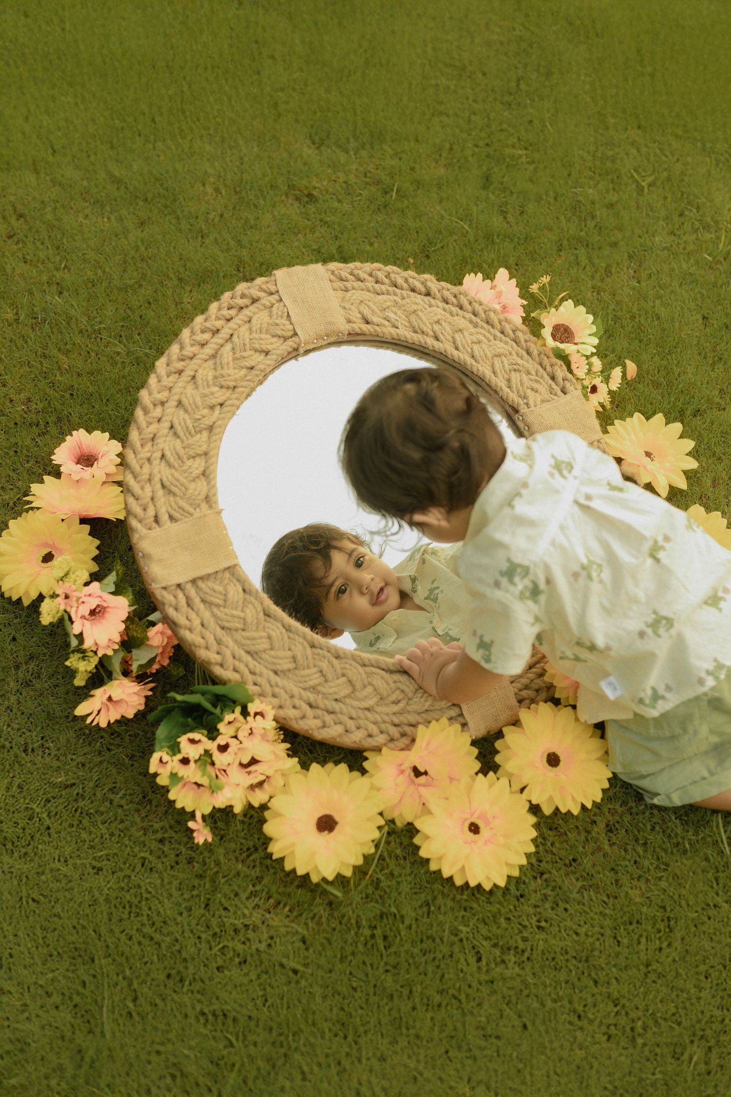 A child rests on a round woven mat surrounded by pink and yellow daisies on a green carpet.