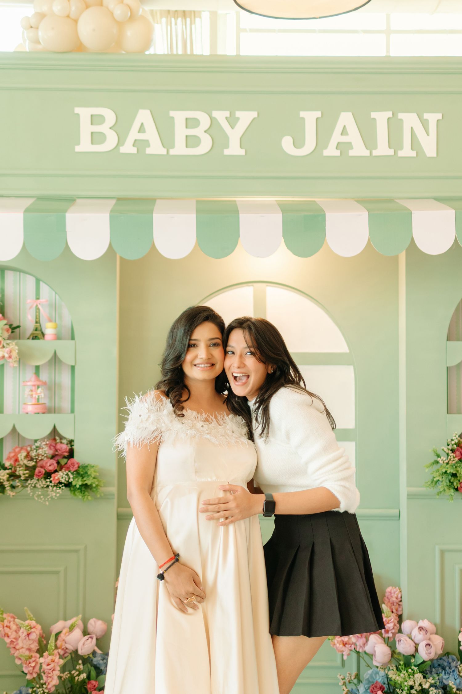 Two women pose together at a Baby Jain baby shower in front of a mint green decorative backdrop with flowers.
