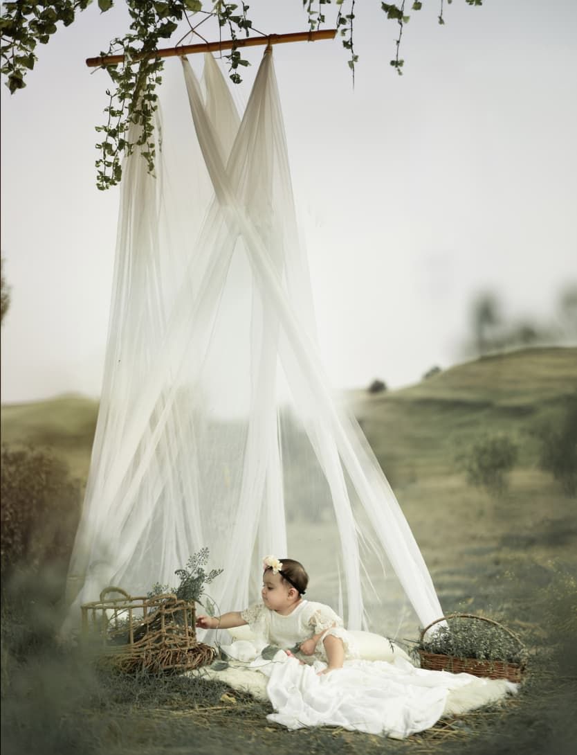 Baby in white dress sits under sheer canopy outdoors on blanket with wicker baskets in dreamy nature setting.