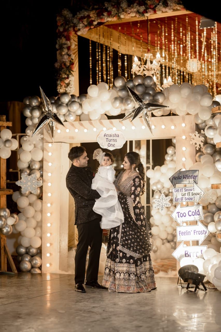 Family celebrating a first birthday party under a decorative balloon arch with winter theme, star ornaments, and string lights.