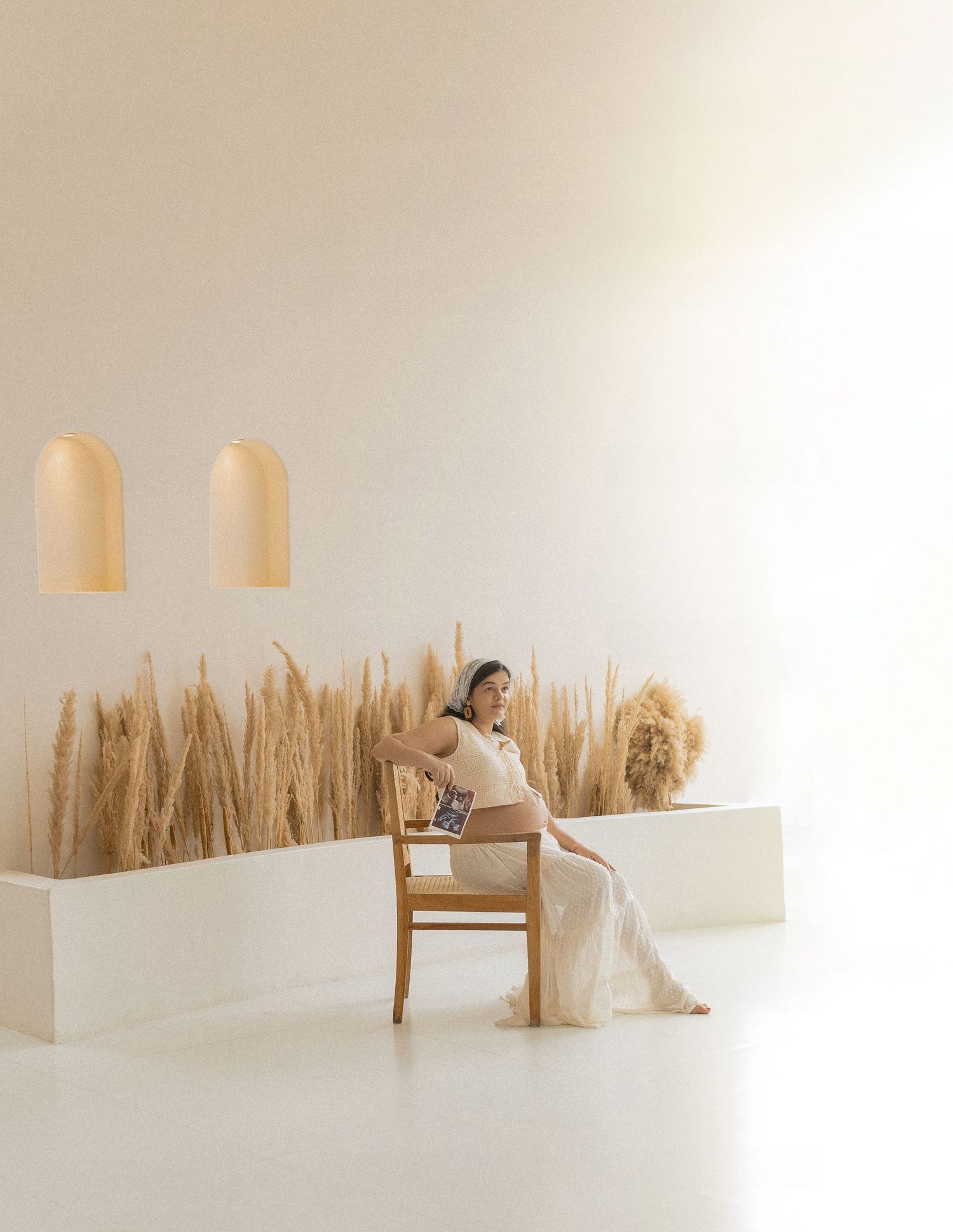 Pregnant woman in white dress sits on wooden chair surrounded by dried pampas grass in minimalist beige studio.