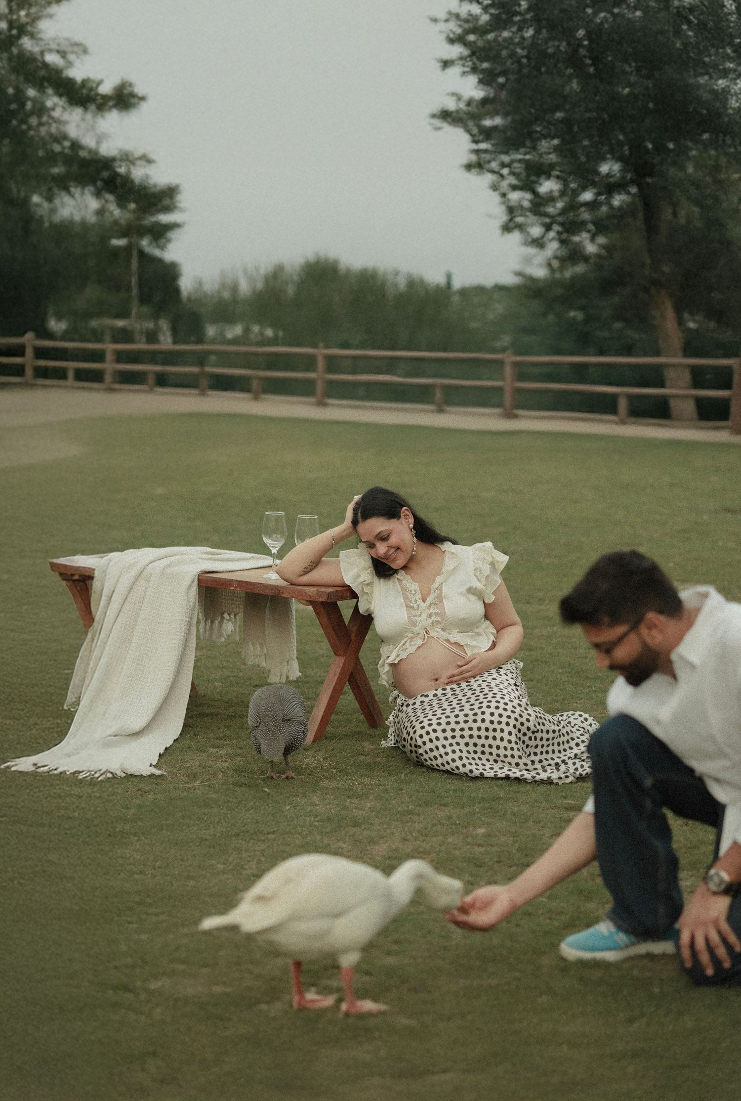 Pregnant woman sits on grass near picnic table while man feeds white duck outdoors in a serene park setting.