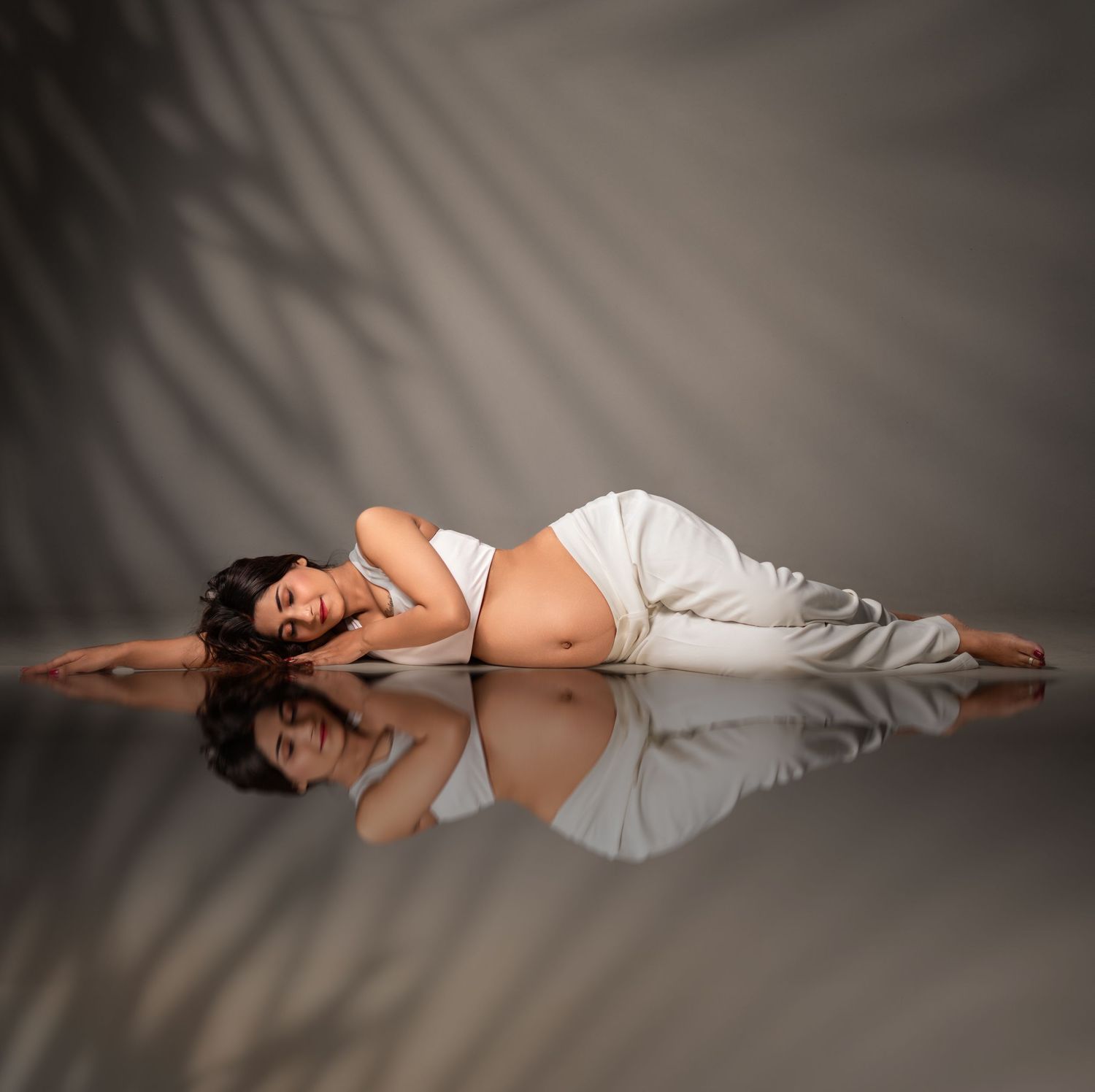 Artistic maternity photo with white fabric on reflective floor and dramatic palm shadow backdrop.