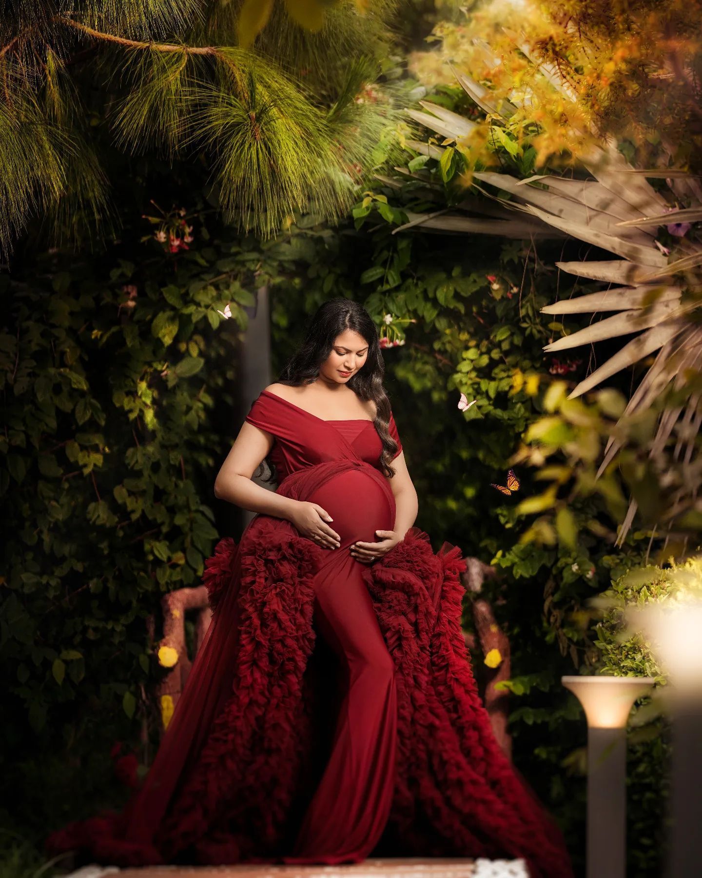 Garden maternity portrait featuring dramatic red off-shoulder gown with ruffled details.