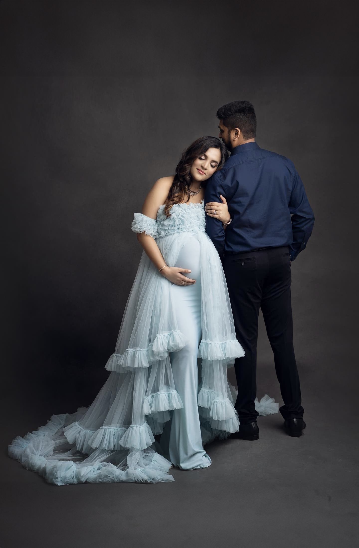 A romantic maternity photoshoot with a flowing light blue tulle gown against a dark backdrop.