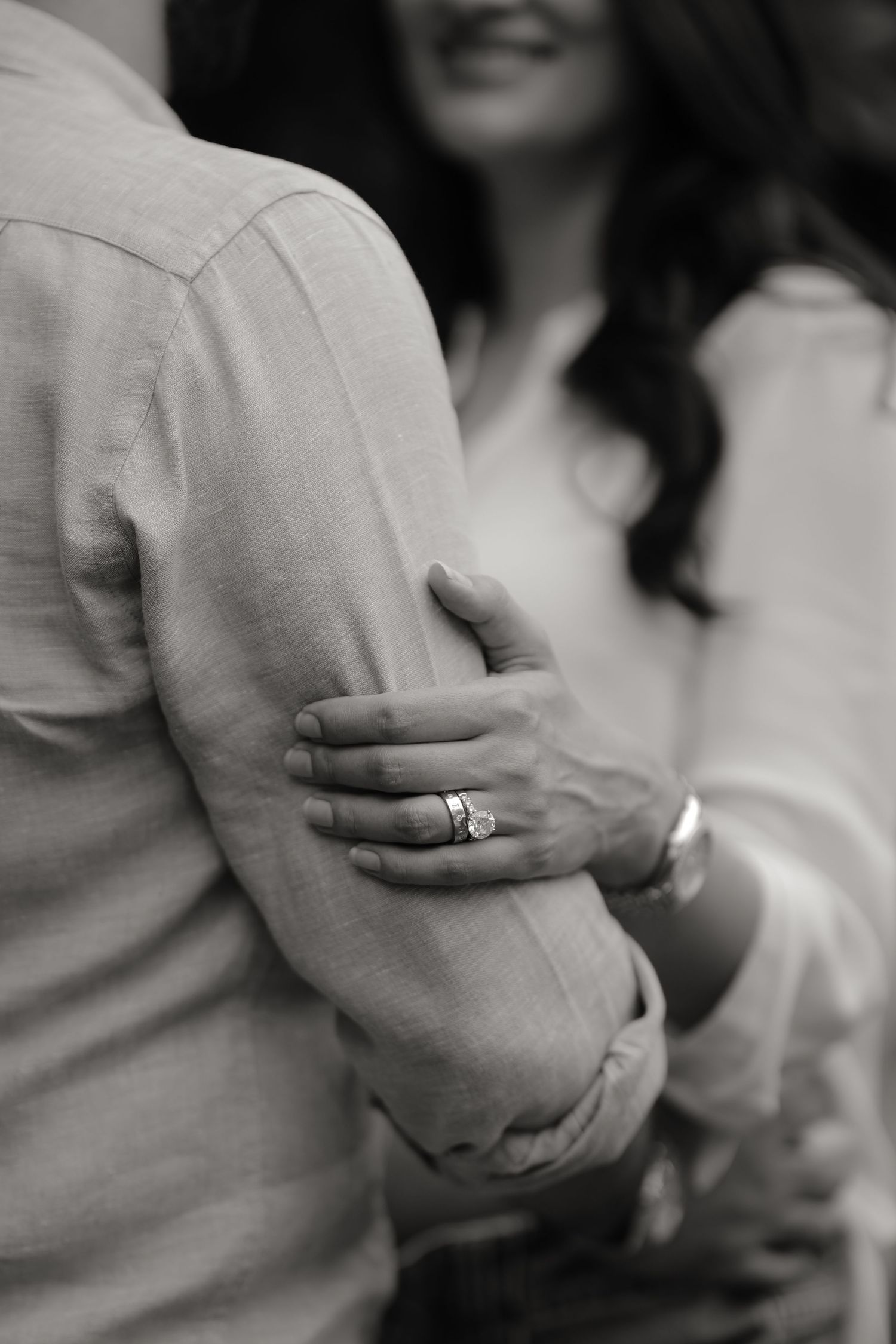 Black and white photo of a couple embracing, woman's hand with engagement ring wrapped around man's arm.