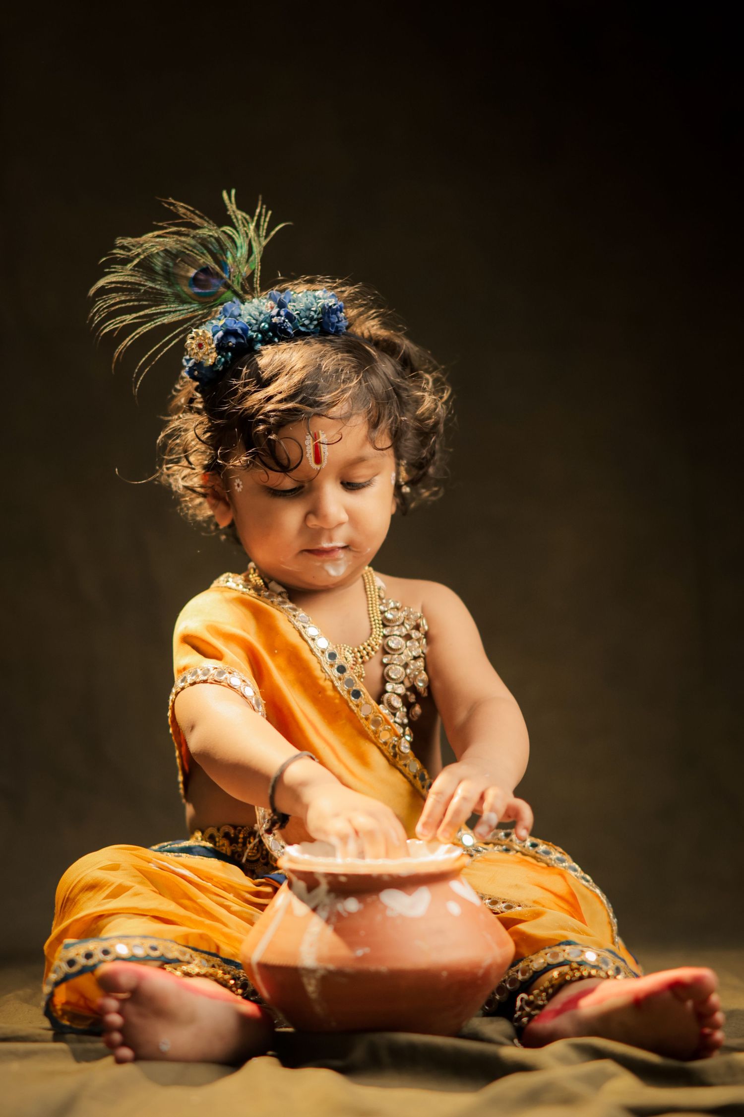 Young child dressed as Lord Krishna in yellow outfit with peacock feather crown, sitting cross-legged with clay pot.