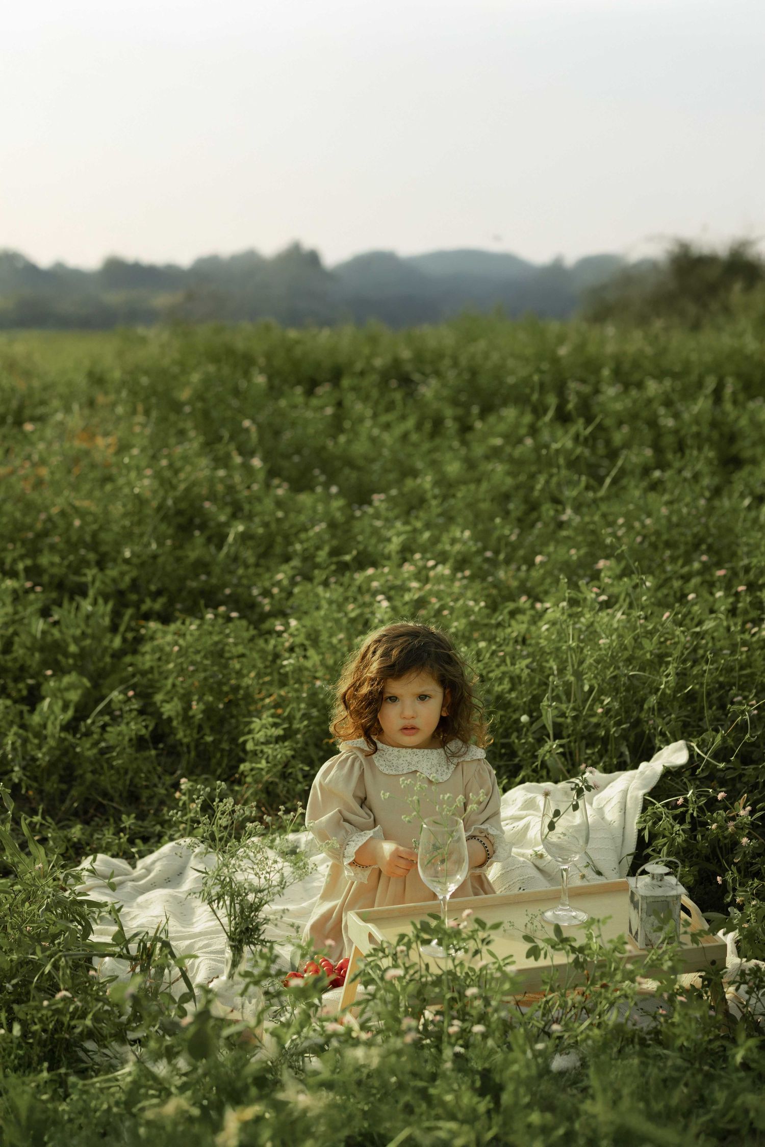 Young girl in white dress sitting on blanket in lush green field for outdoor picnic.