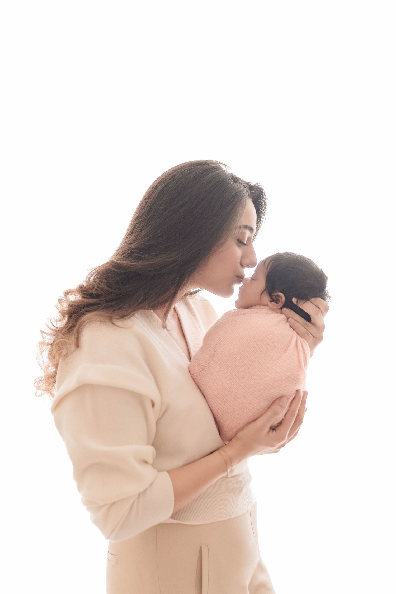 A tender moment as a young mother in beige holds and kisses her newborn baby wrapped in a pink blanket.
