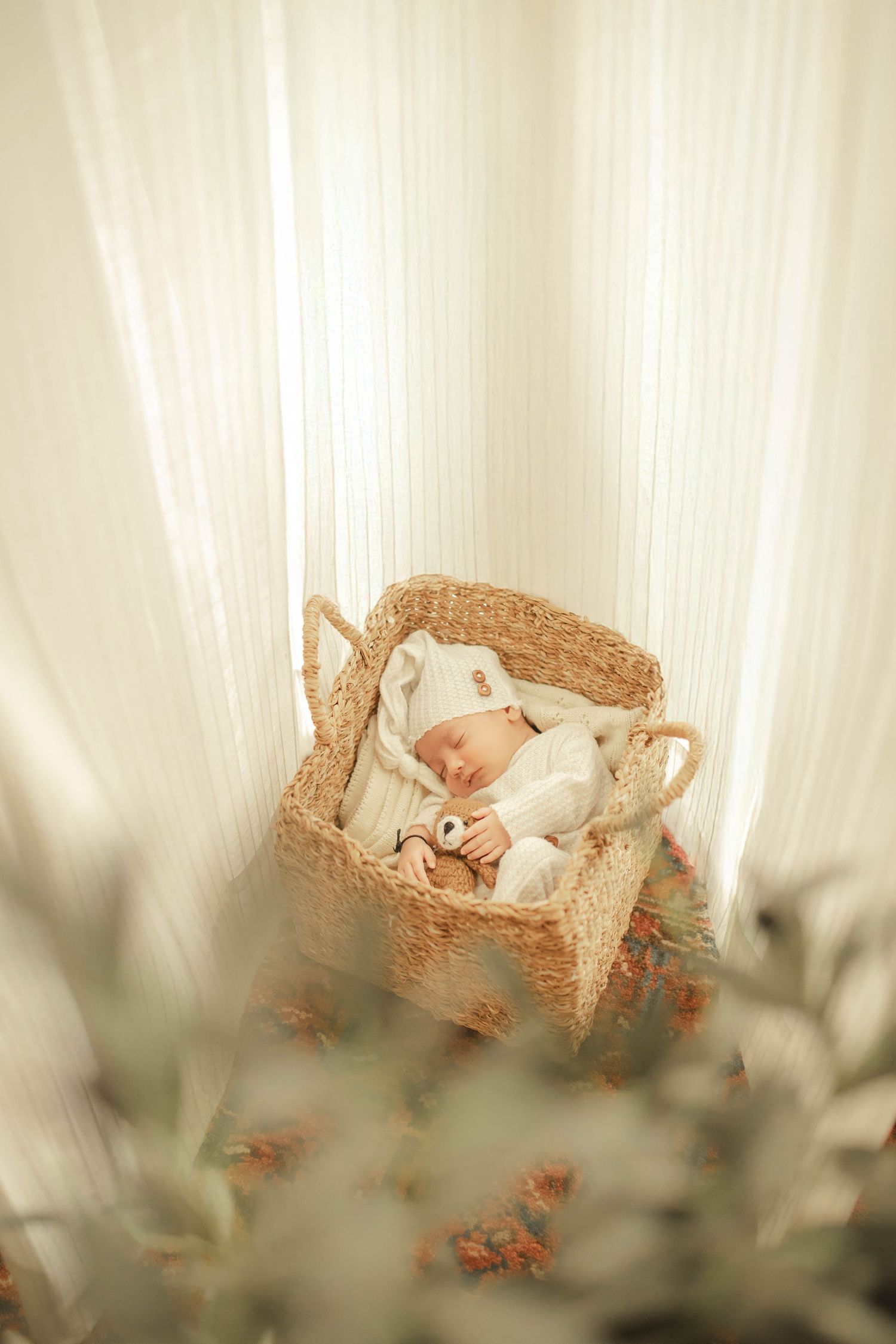 Newborn baby sleeping peacefully in a wicker Moses basket, dressed in white outfit and hat, surrounded by soft curtains.