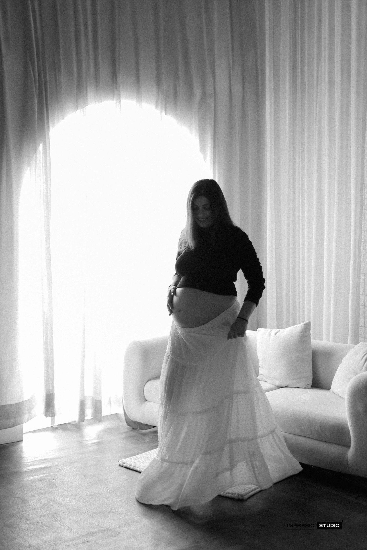 Black and white maternity photo of pregnant woman in black top and white skirt standing by bright arched window.