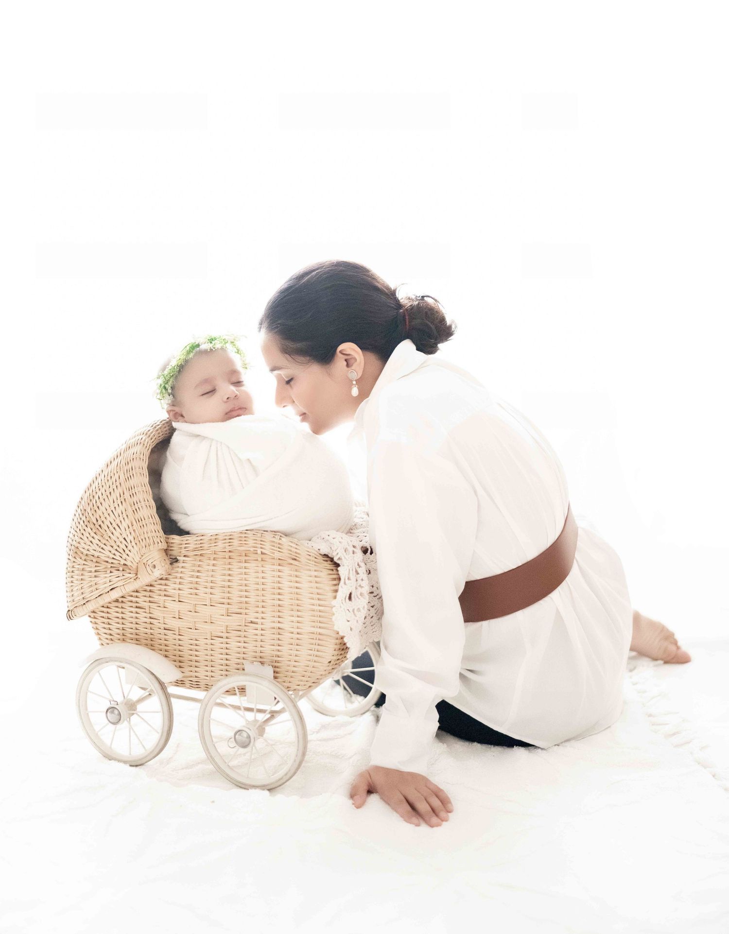 Mother in white dress kisses newborn baby with flower crown in wicker pram on white background.