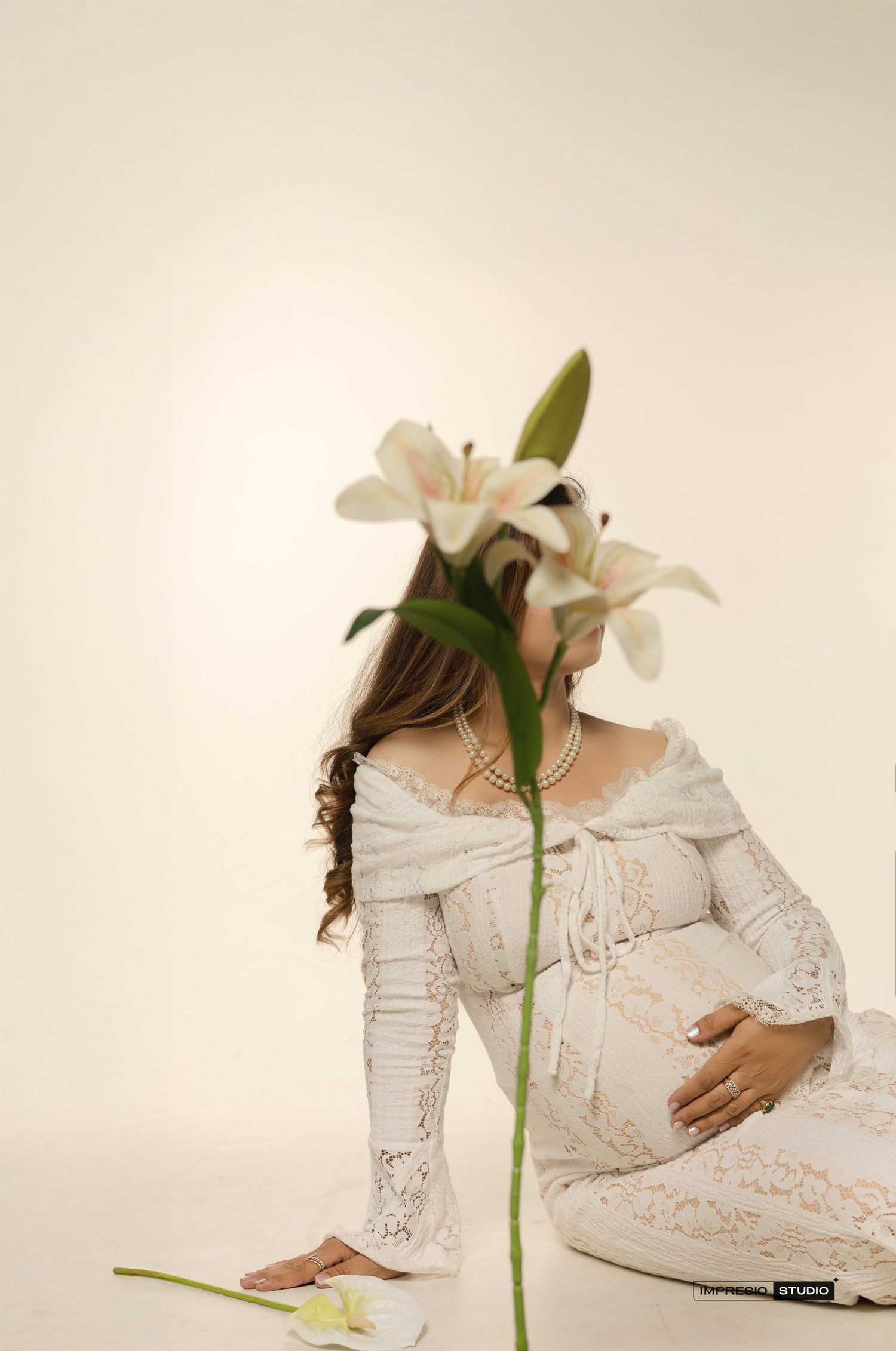Pregnant woman in white lace off-shoulder gown sits gracefully beside white lilies in a soft studio setting.