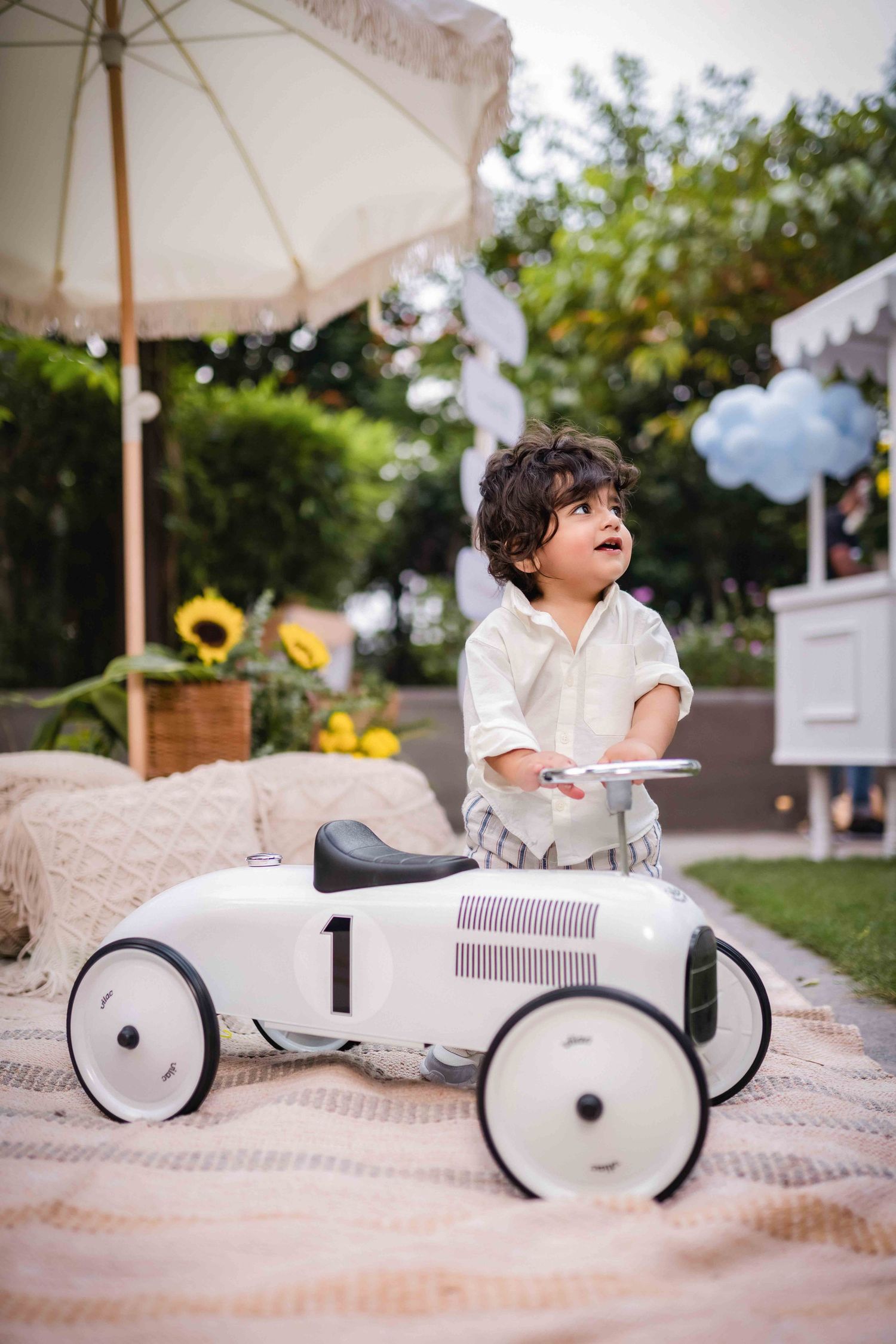 A toddler boy sits in a white vintage toy race car numbered 1 outdoors at a garden party with balloons.