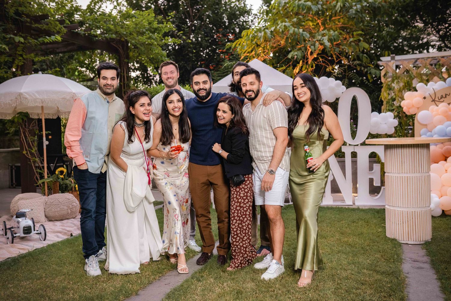 Group of friends posing together at an outdoor garden party with balloon decorations and festive signage.