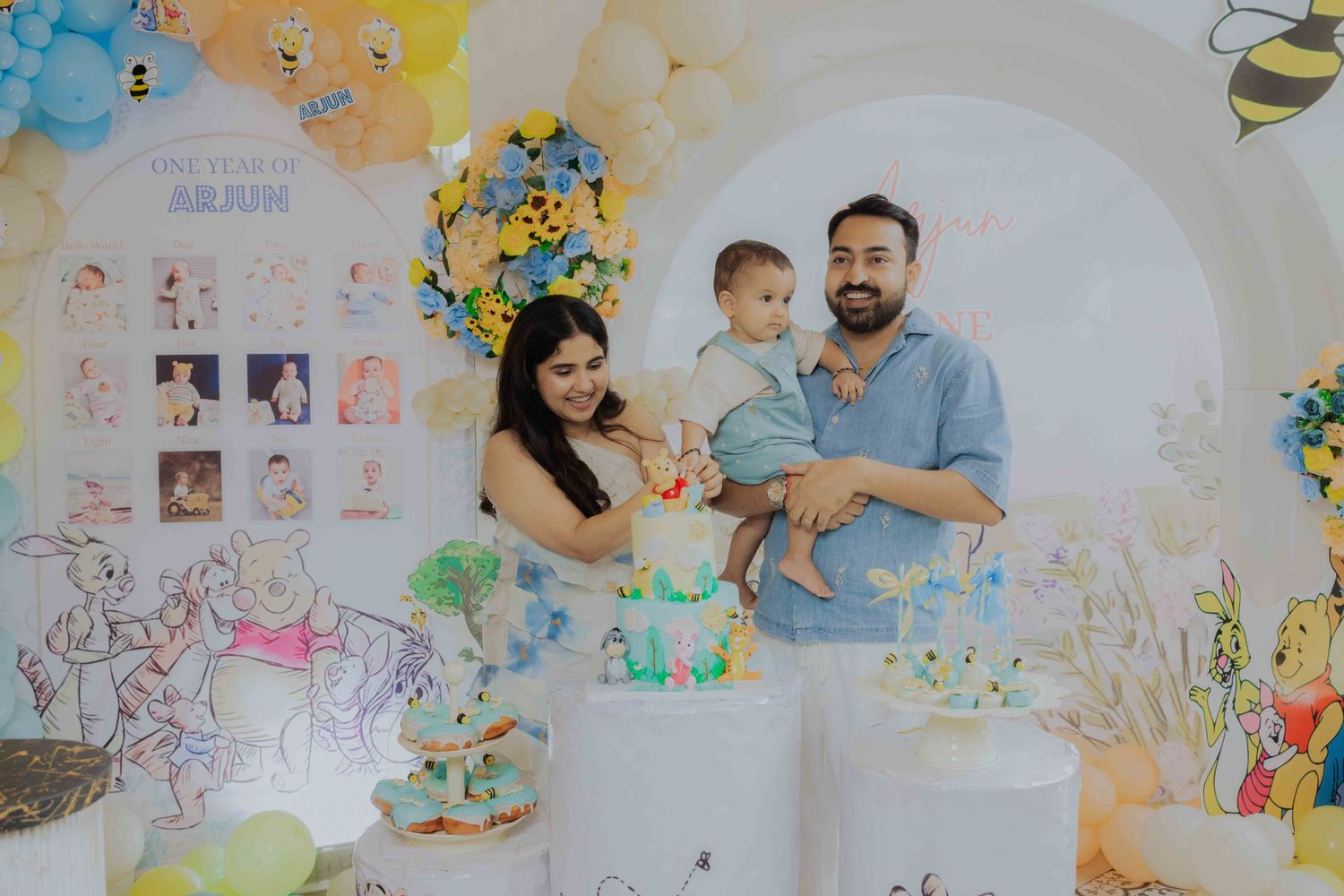 Family celebrating baby's first birthday with Winnie the Pooh themed cake and balloon decor.