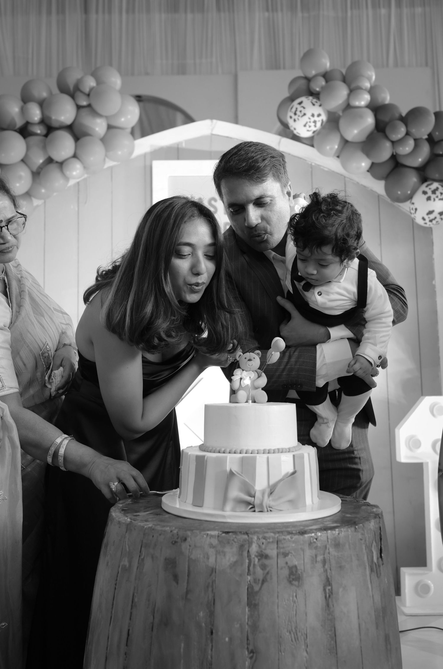Black and white photo of a family celebrating a baby's first birthday, cutting a two-tier cake with balloon decorations.