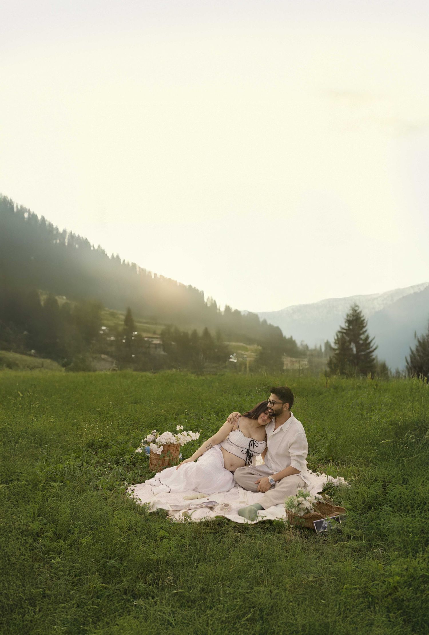 Couple sitting on blanket in green mountain meadow at golden hour, woman in white dress, romantic maternity photoshoot.