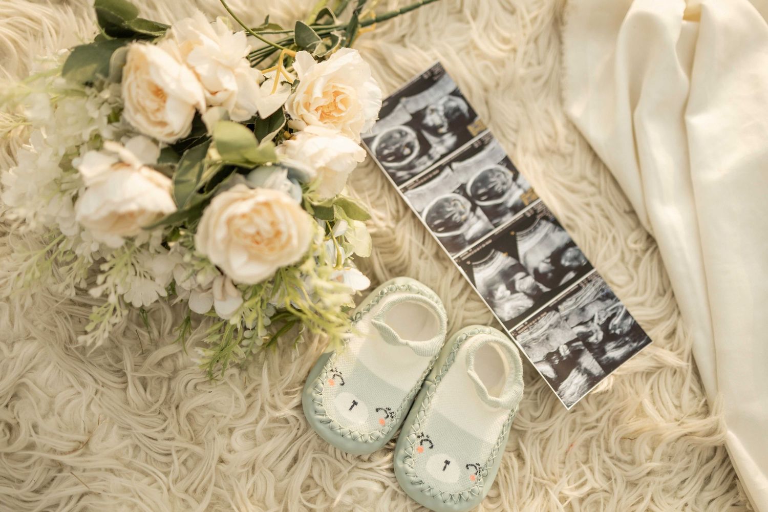 Baby shoes, ultrasound photos, and white roses bouquet arranged on a soft cream fur blanket for a pregnancy announcement.