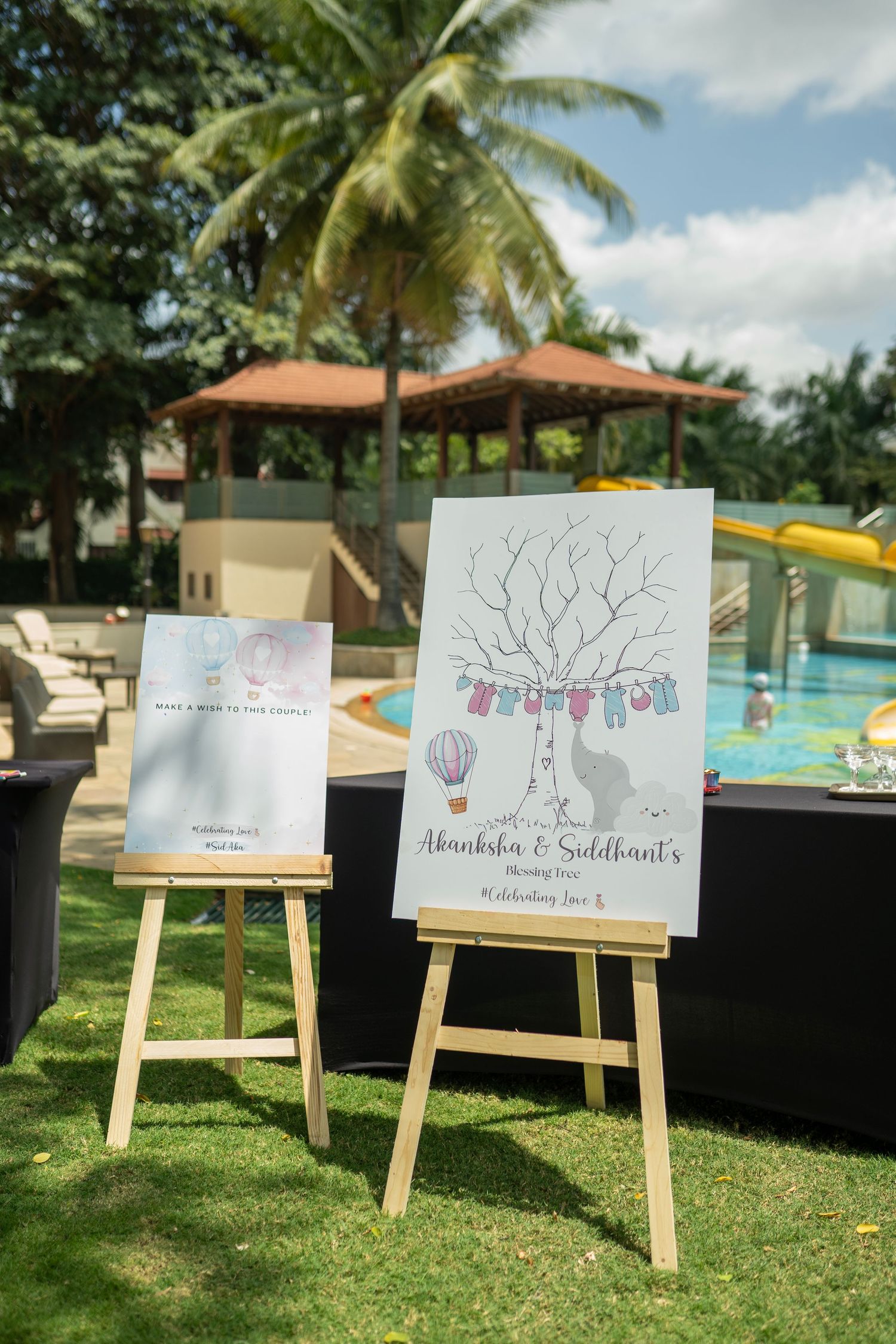 Outdoor wedding welcome signs on wooden easels by a pool, featuring a fingerprint tree design for Akanksha & Siddhant.
