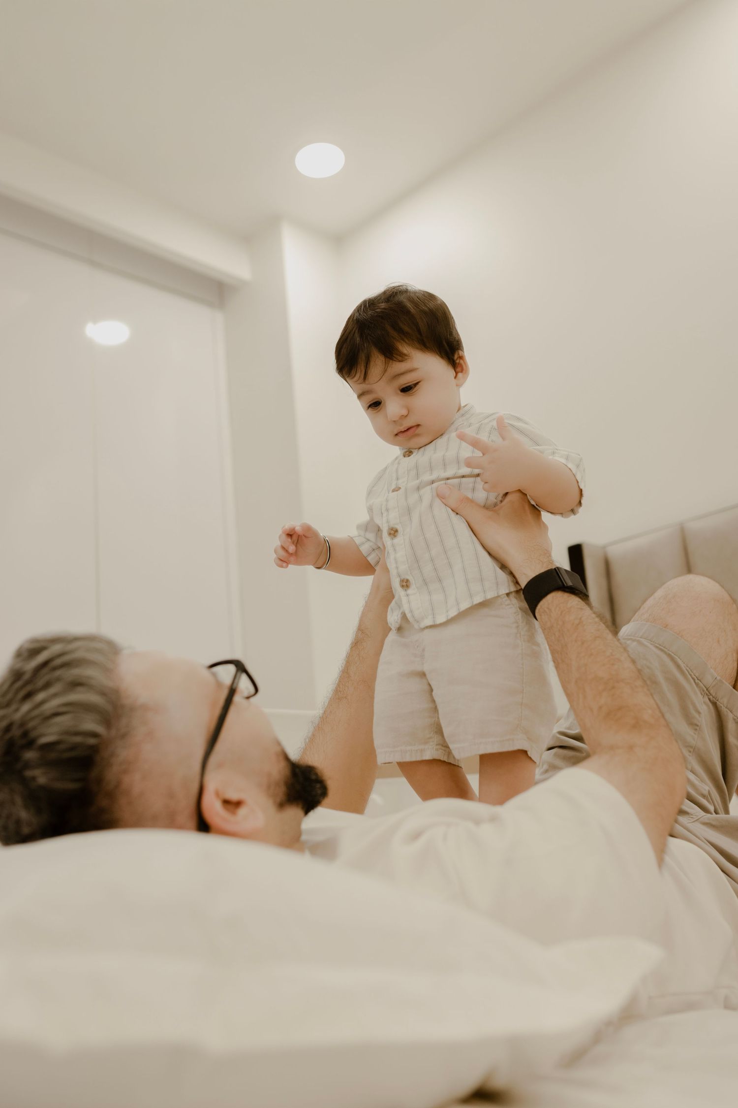 A parent plays with a baby wearing white clothes on a light-colored bed in a bright white room.